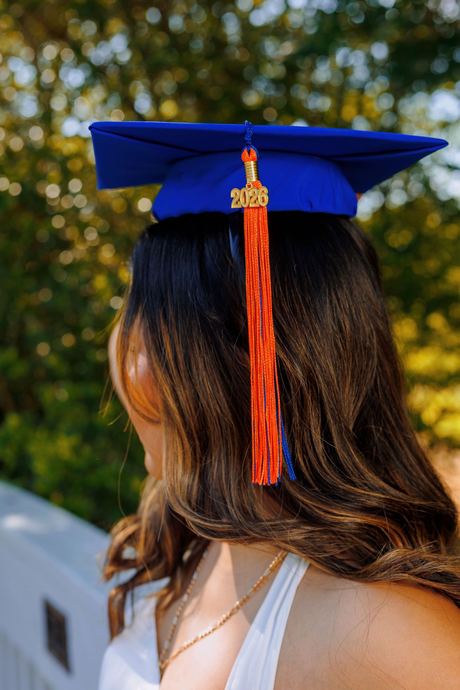 Back view of graduate wearing blue 2026 mortarboard cap with orange tassel among green trees.