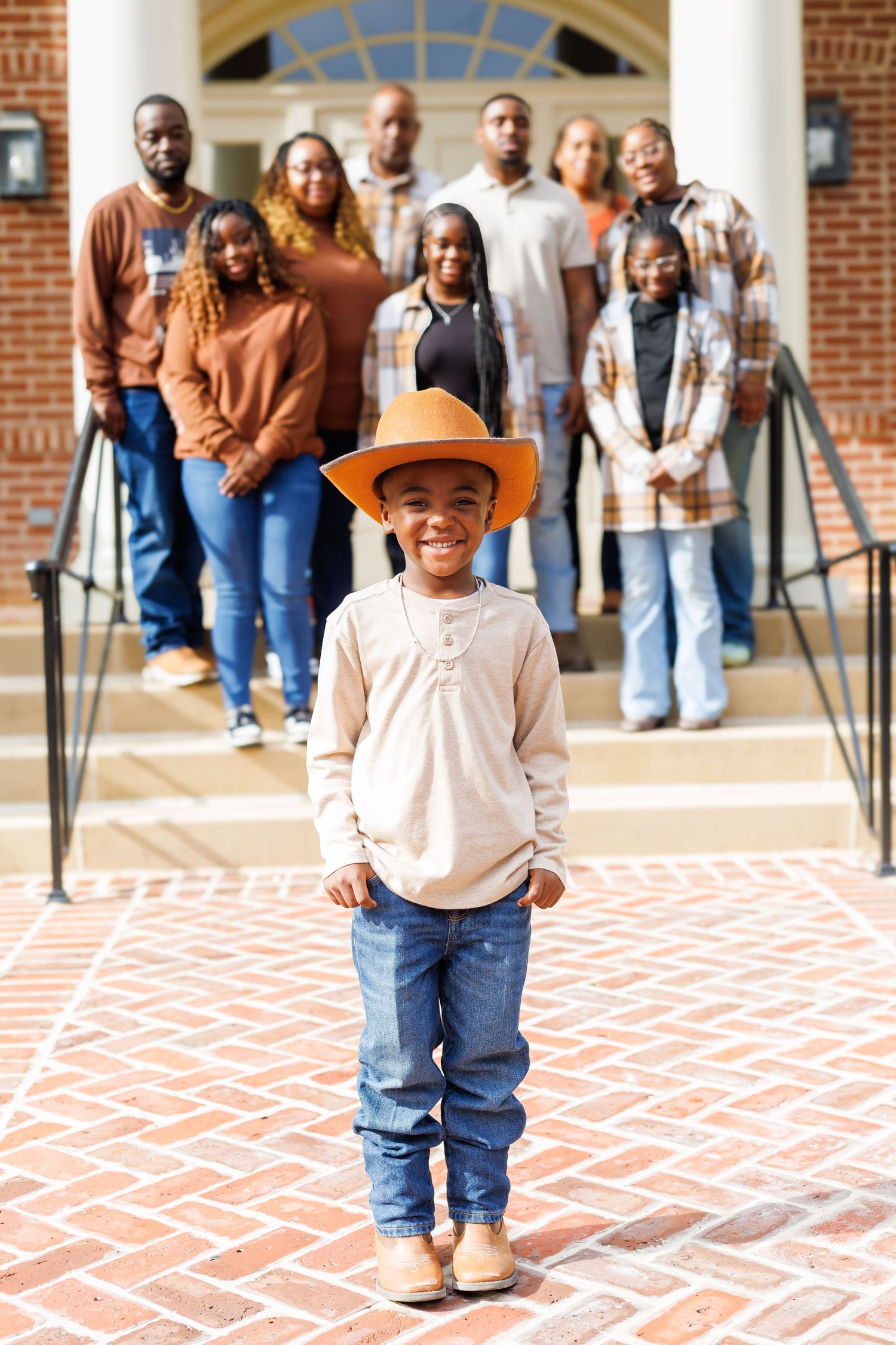 Smiling young boy in cowboy hat and jeans standing before a large family group on brick steps.