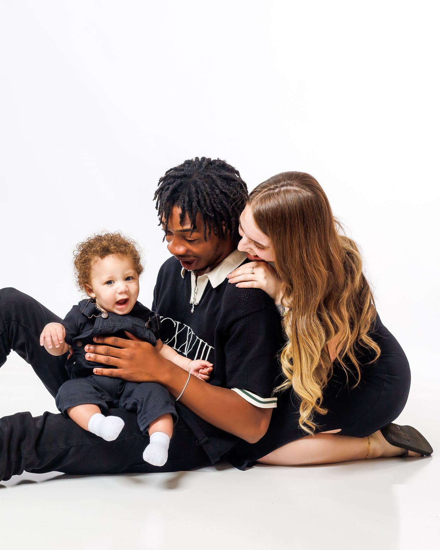 A young family sits together on a white background, parents smiling at their happy baby dressed in black.