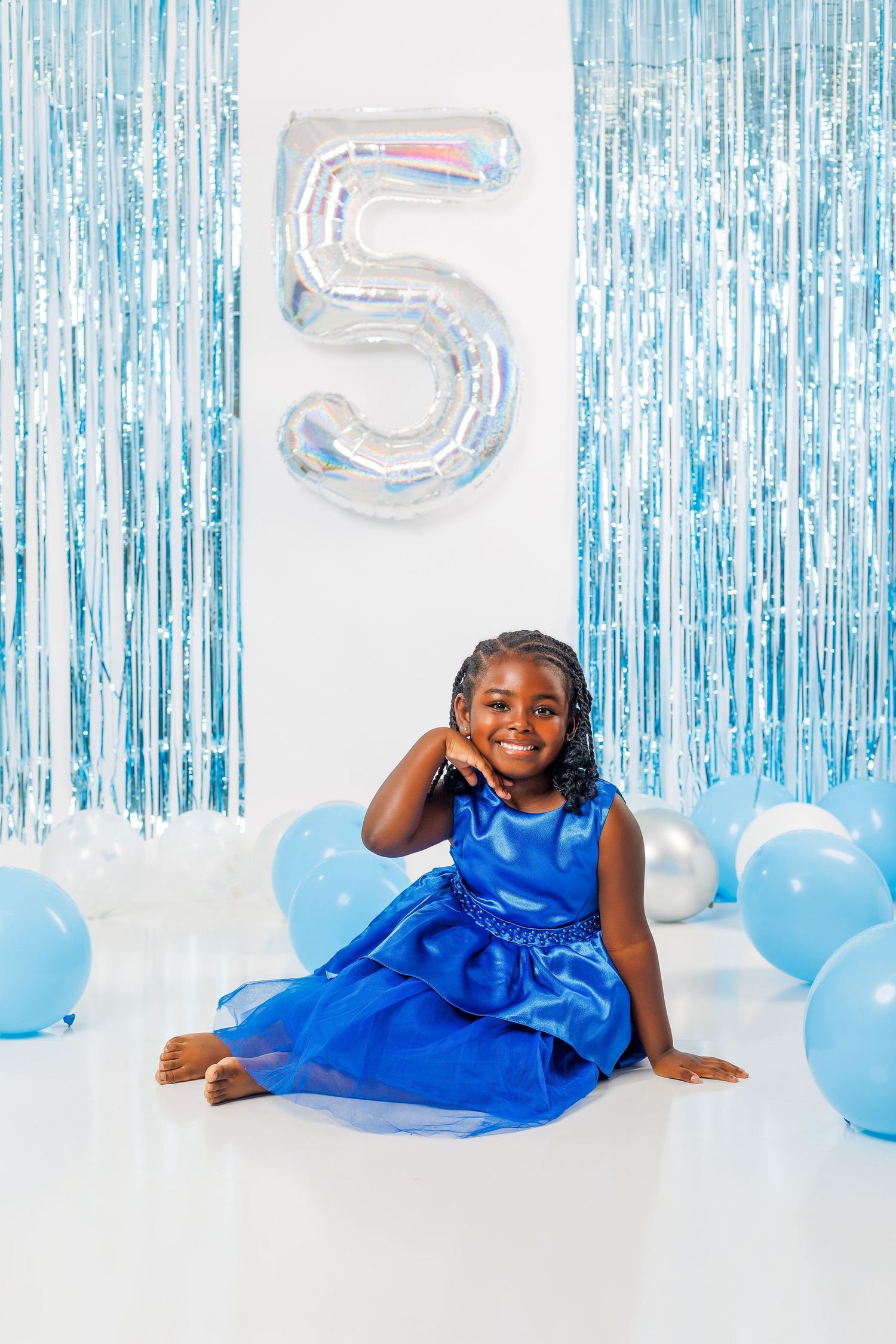 Happy girl in blue dress sits on floor smiling beside silver number 5 balloon with blue party decorations.
