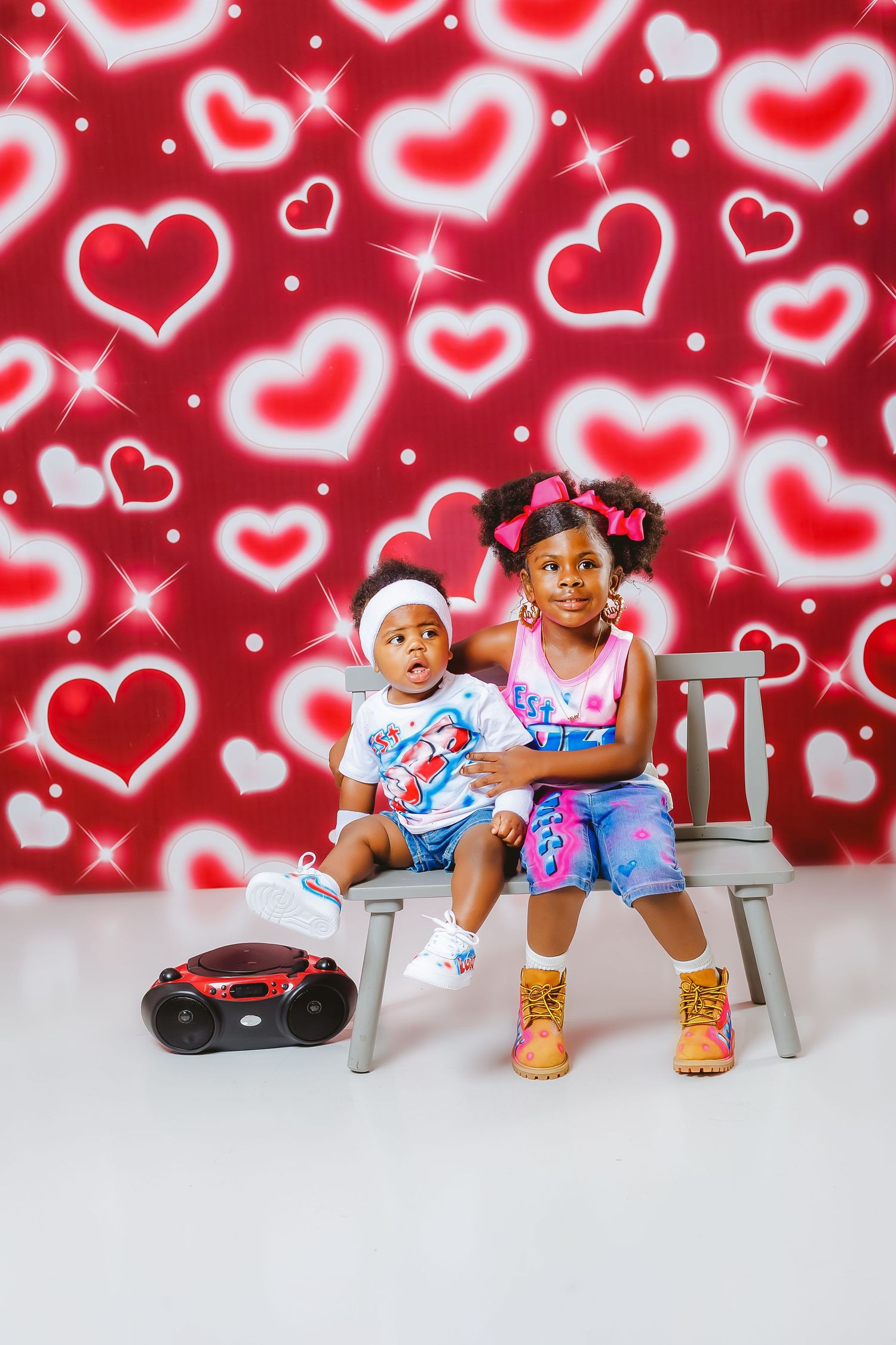 Two young sisters sit together on bench in front of red heart-patterned backdrop with boombox beside them.