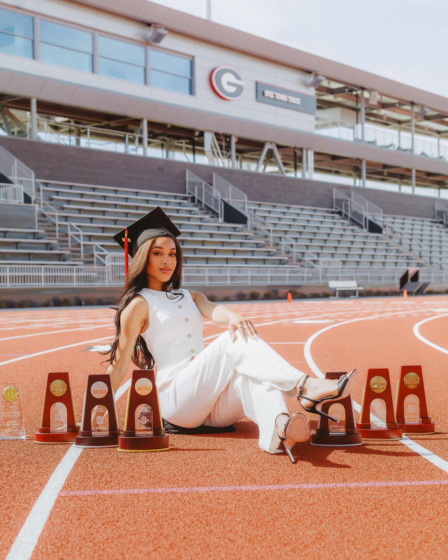 Female graduate in white outfit sitting on track field surrounded by NCAA championship trophies.