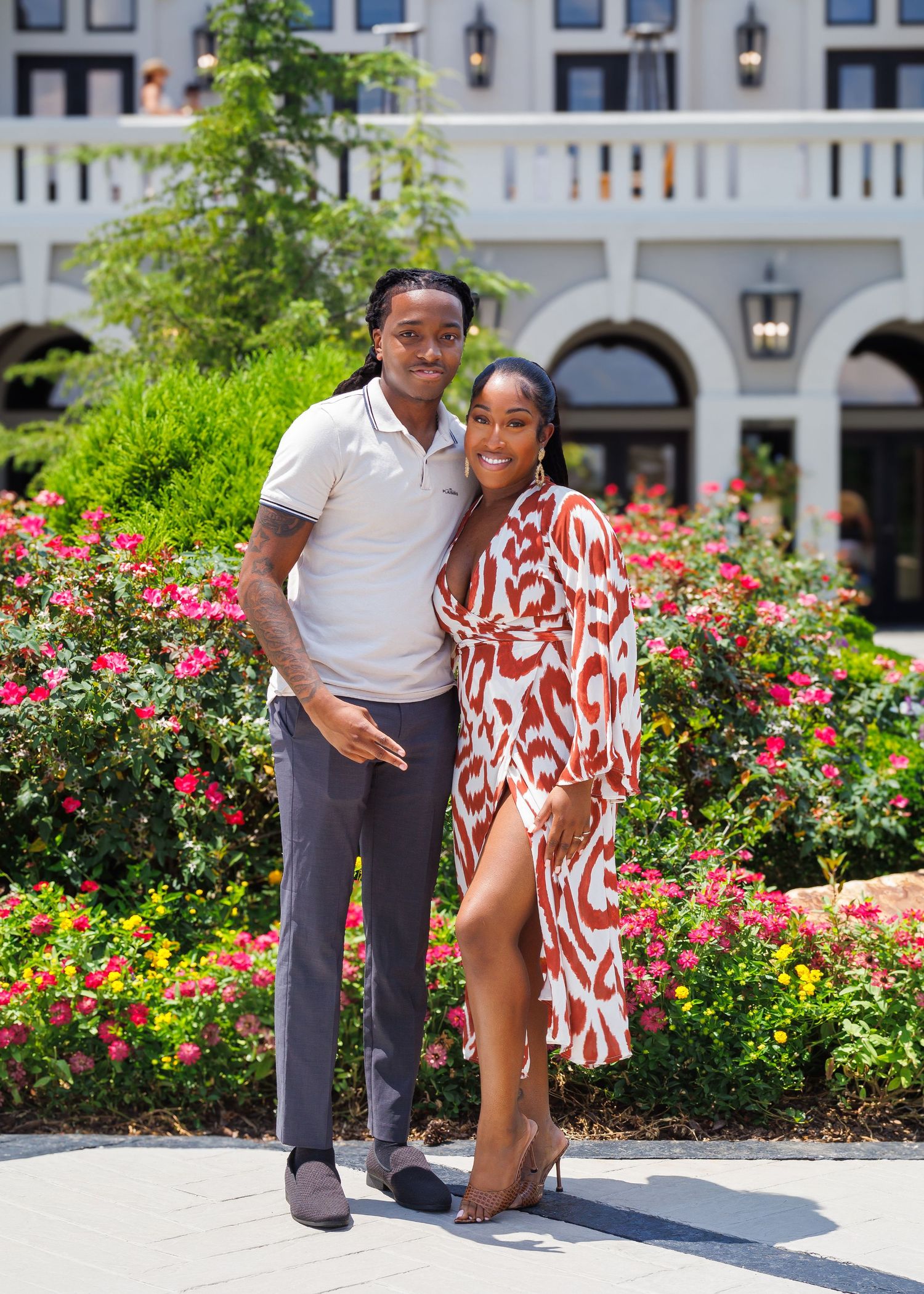 Couple posing together in front of colorful flower garden outside elegant building on sunny day.
