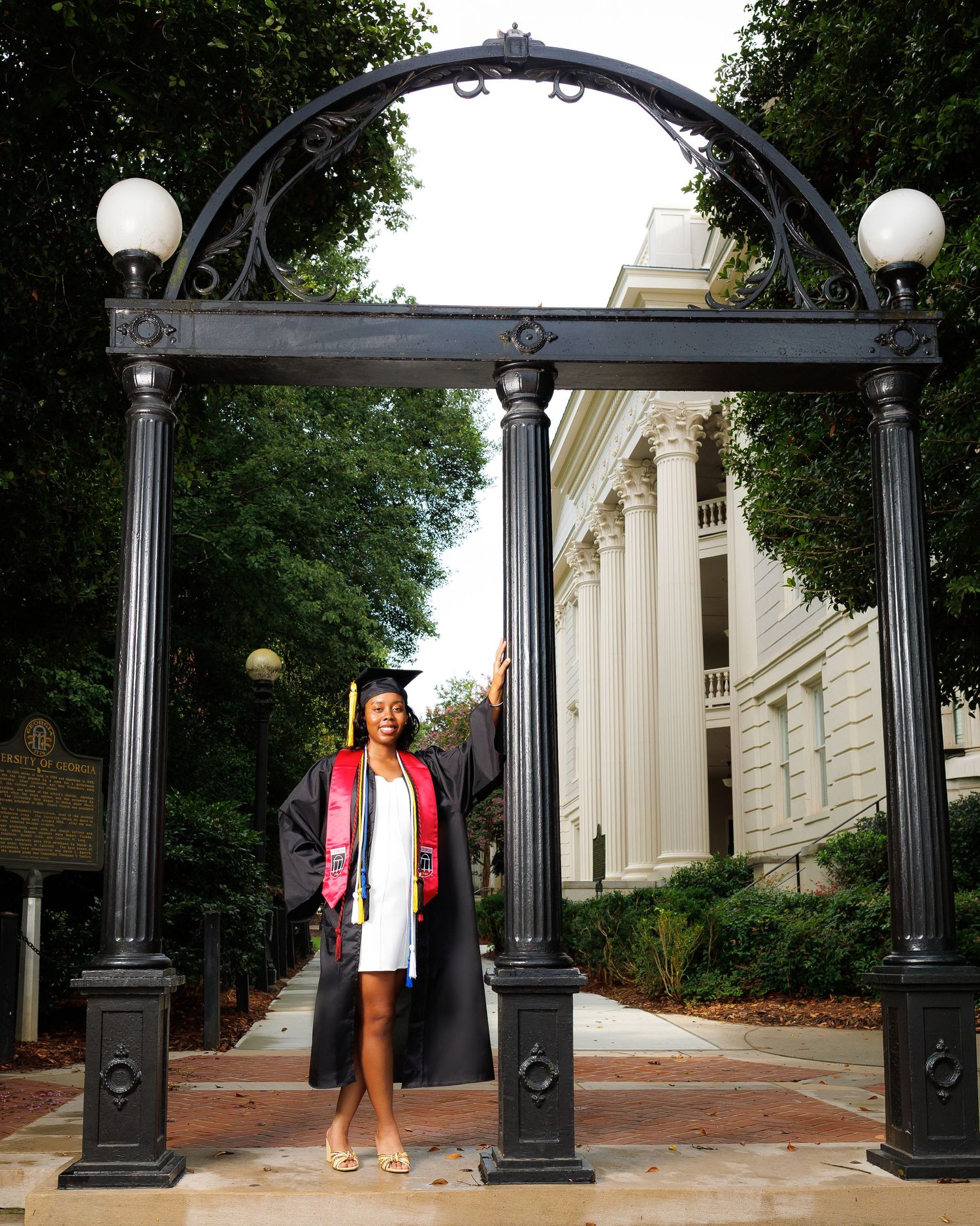 Graduate in cap and gown posing under the iconic University of Georgia arch landmark.