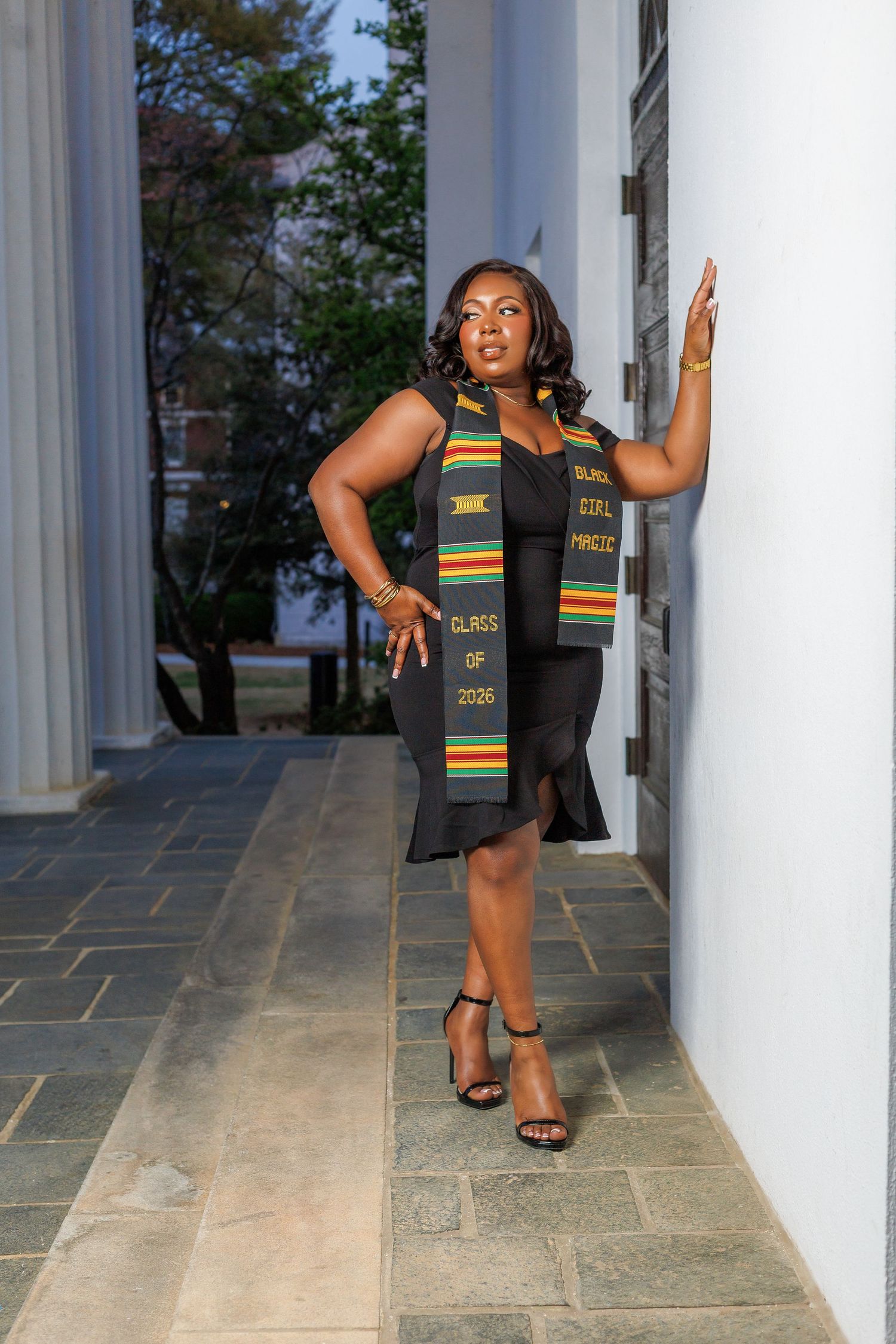 Graduate posing against white column wearing Black Girl Magic Class of 2020 Kente graduation stole.