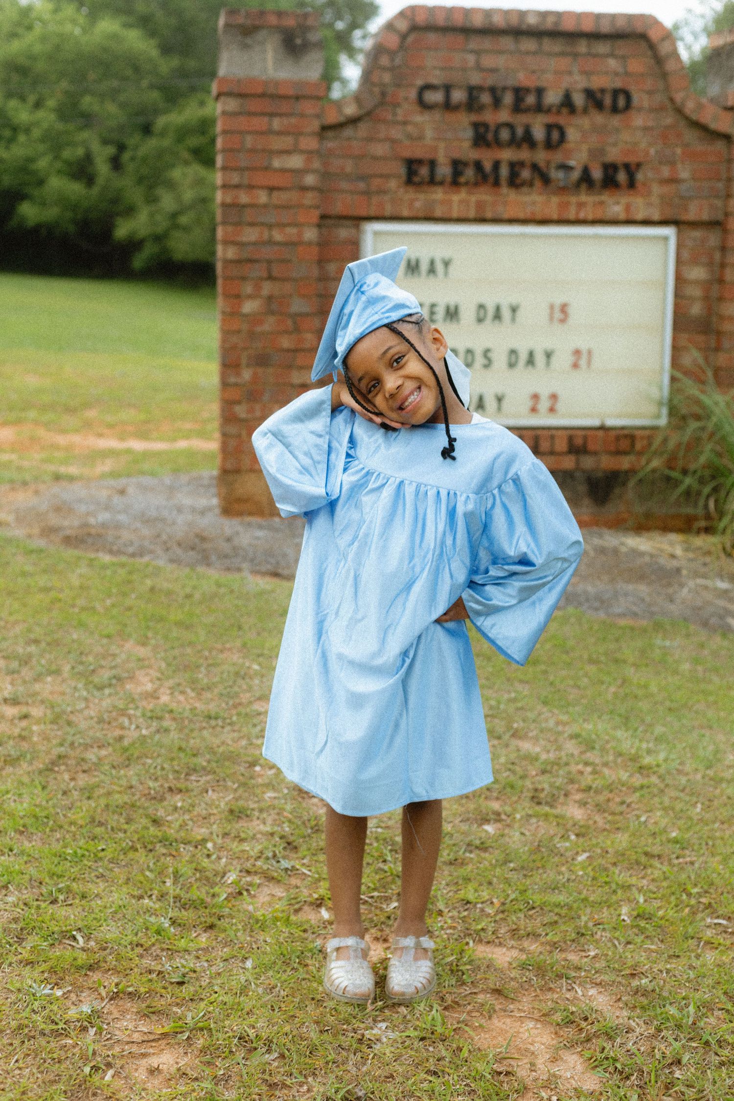 Young girl in light blue graduation cap and gown poses in front of Cleveland Road Elementary school sign.