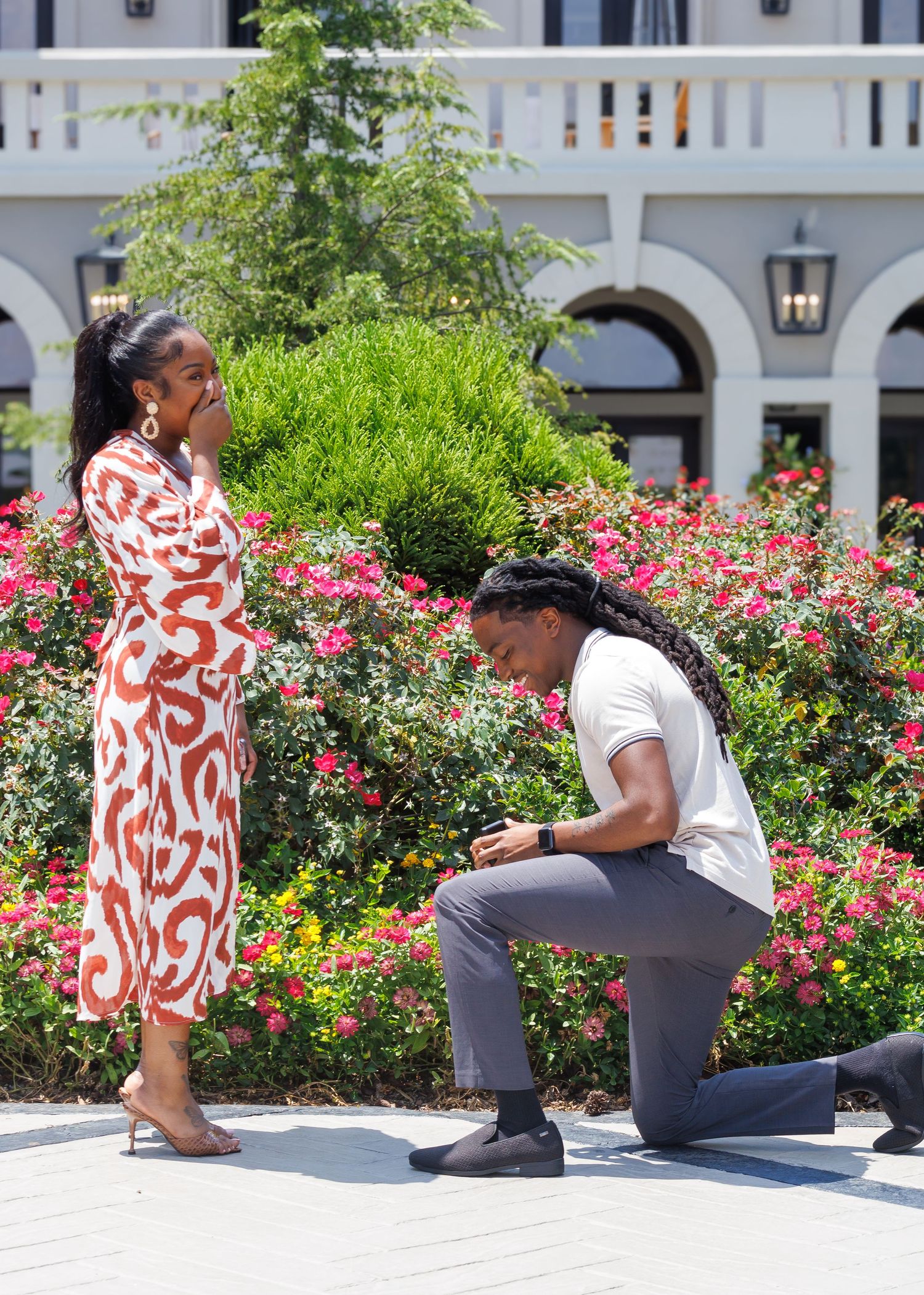 Man kneeling on one knee proposing to woman in red patterned dress in a colorful garden.