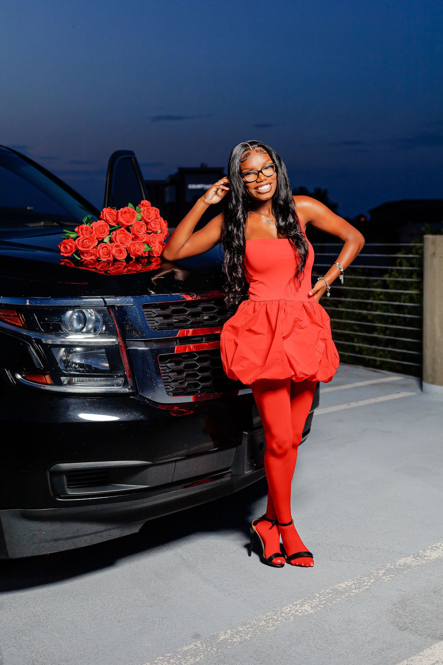 Black woman in red bubble dress and tights poses beside black SUV with red roses bouquet at night.