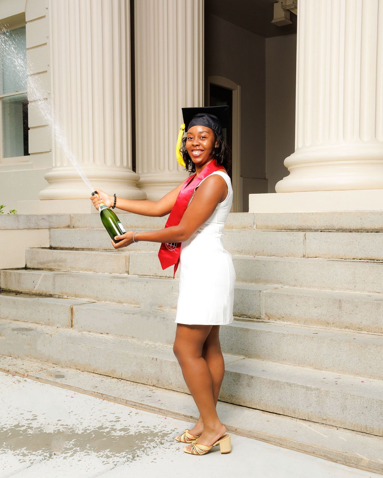 Joyful graduate spraying champagne on university steps wearing white dress and graduation cap.