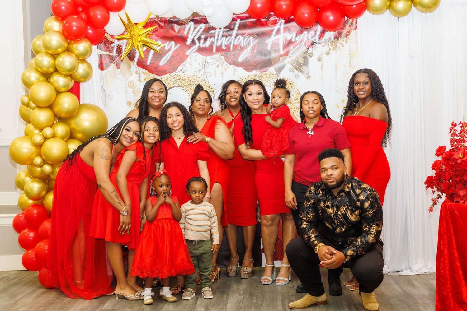 Group of women in red dresses celebrating a birthday party with gold and red balloon decorations.