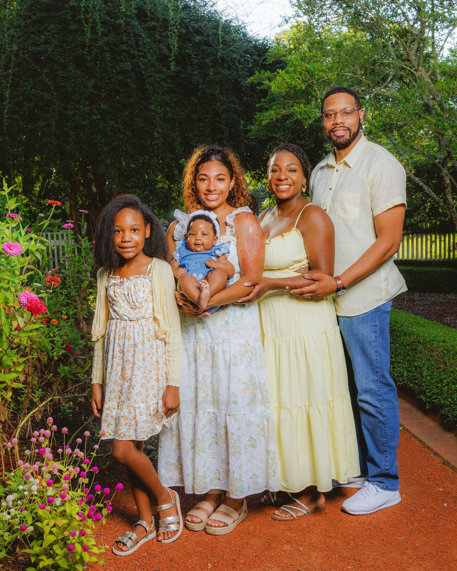 A joyful family of five poses outdoors in a lush garden, dressed in coordinating outfits, holding a baby.