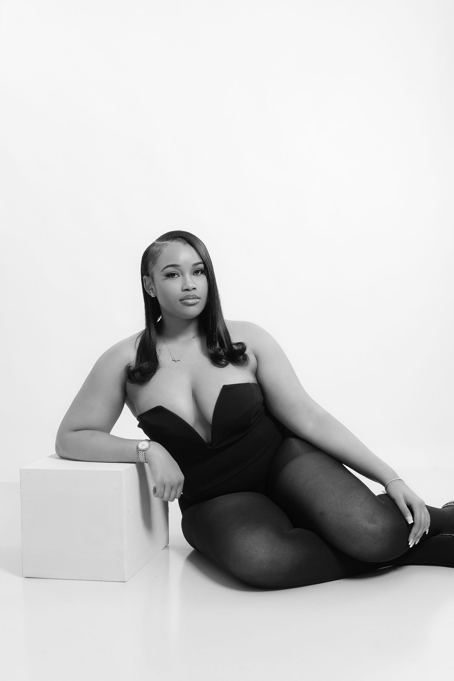 Black and white studio photo of a woman in a black bodysuit sitting gracefully on the floor against a white background.