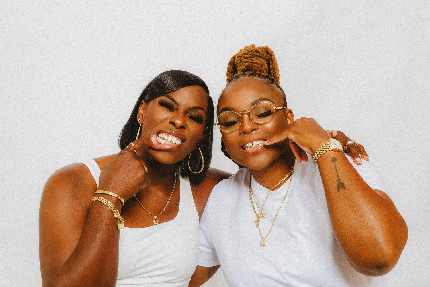 Two smiling women in white tops and gold jewelry posing playfully together.