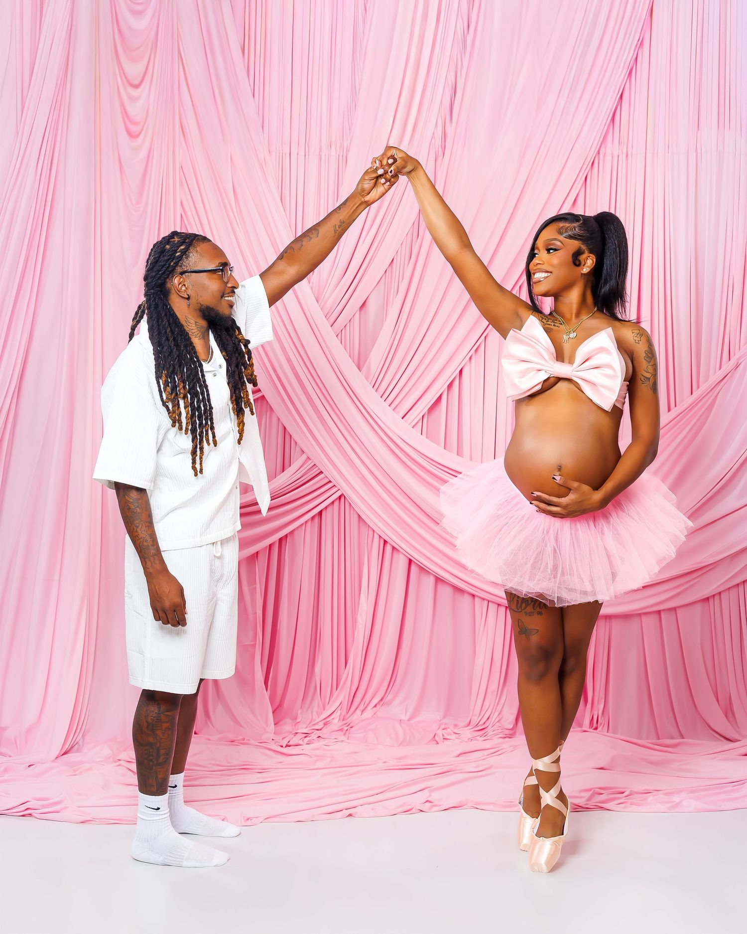 A couple poses together in a pink ballet-themed maternity shoot, with the woman in a tutu.