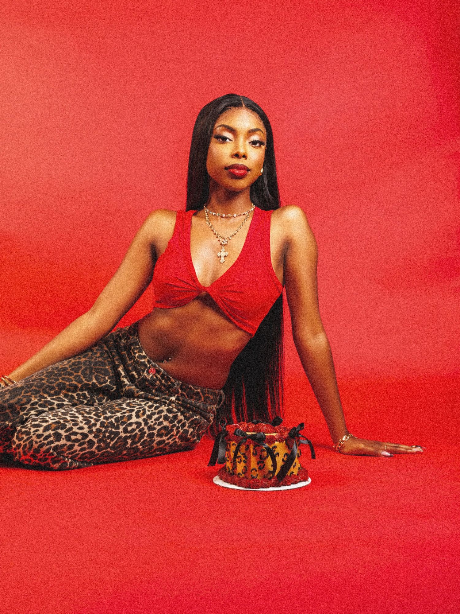 Black woman in red crop top and leopard pants poses with birthday cake against vibrant red background.