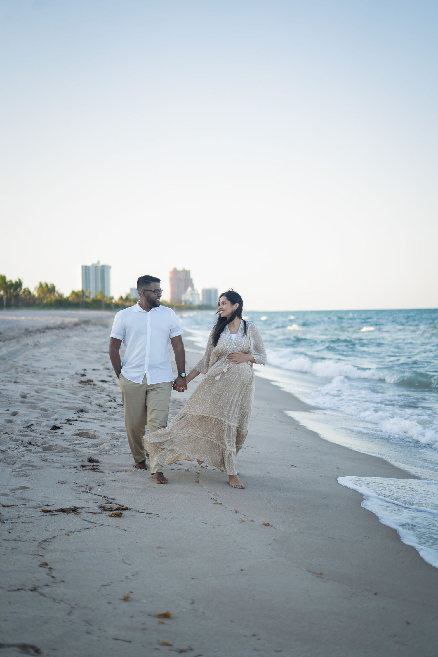 A couple strolls along a scenic beach shoreline with ocean waves and city skyline in the background.