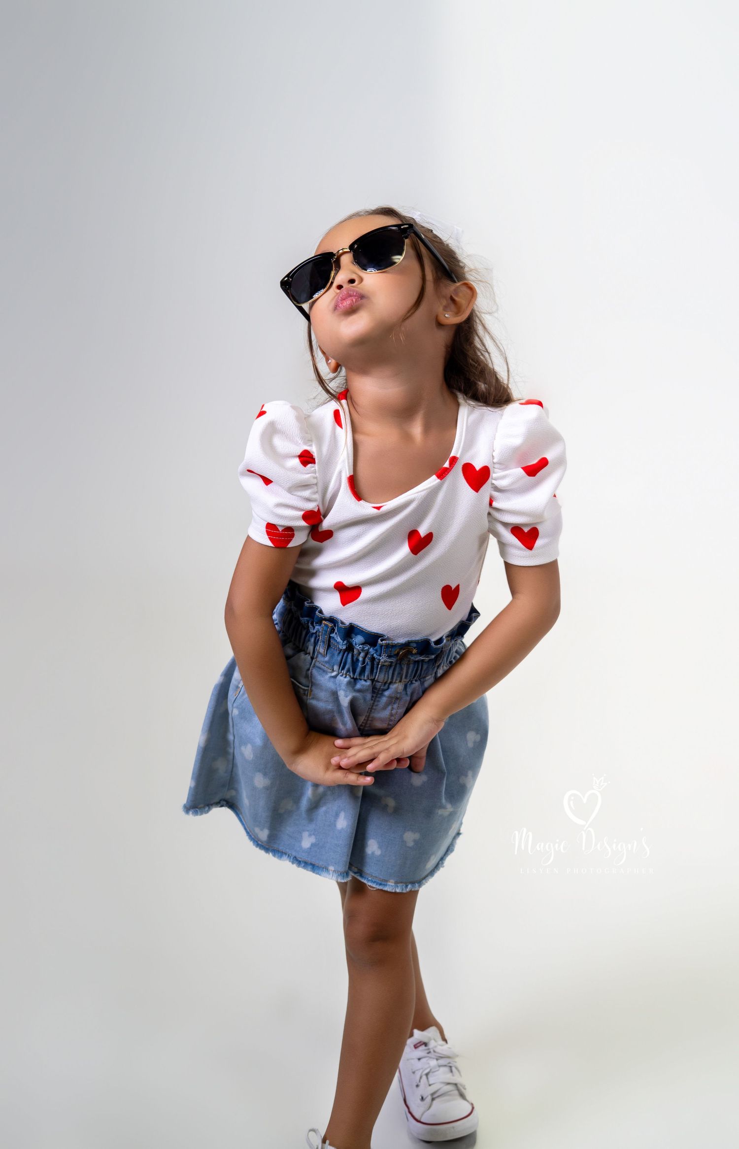 Young child in white shirt with hearts and denim outfit poses playfully in a studio setting.