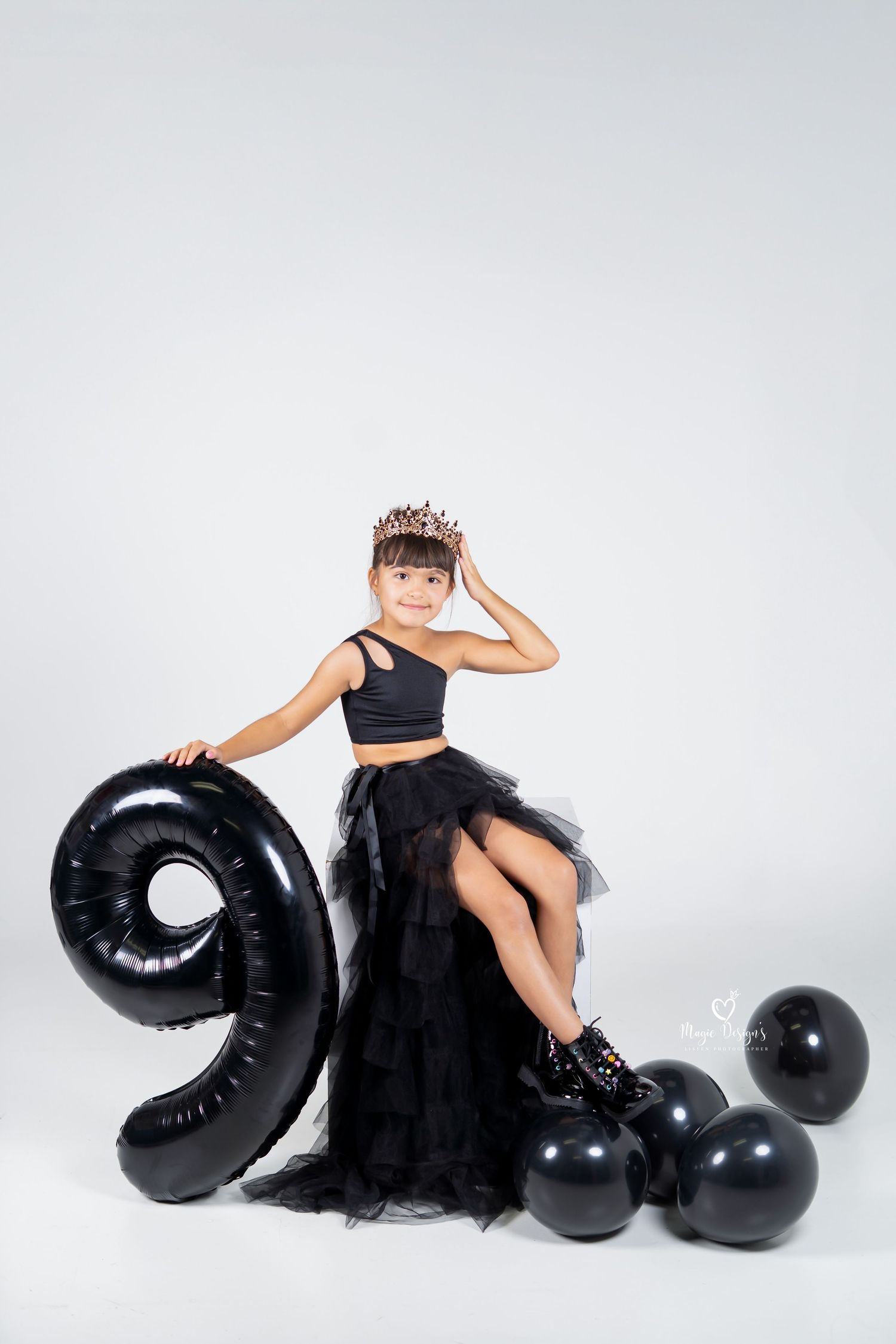 A person in a black tulle dress poses elegantly with large black balloon numbers against a white backdrop.