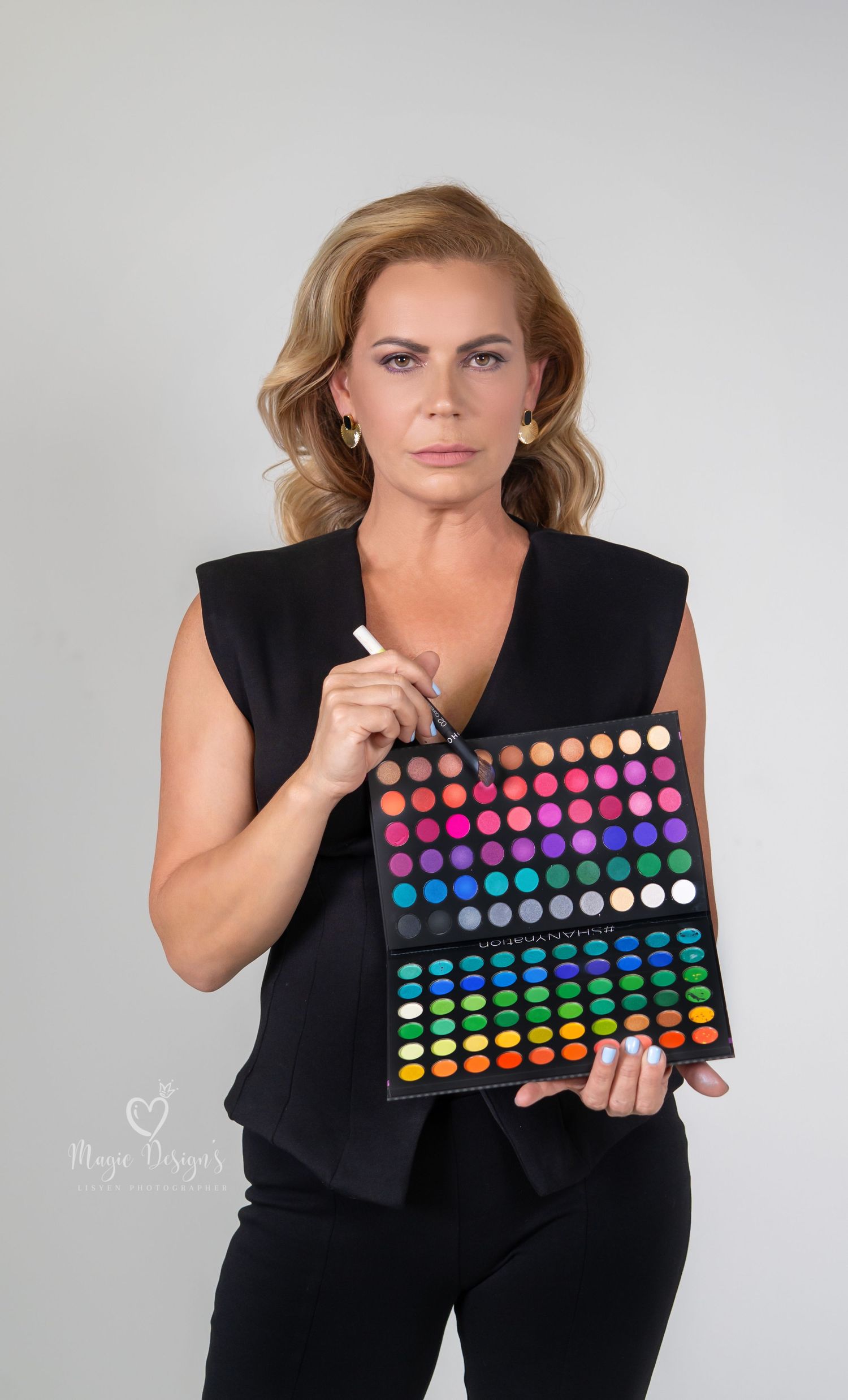 Woman in black outfit displays colorful makeup palette against white studio background.