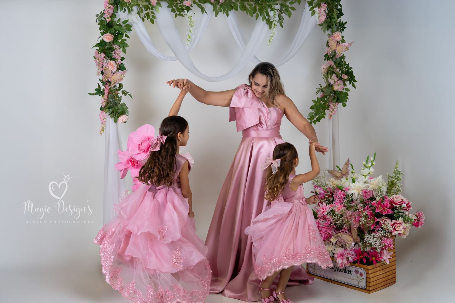 Joyful dance pose in matching pink gowns surrounded by floral decorations and white drapery.