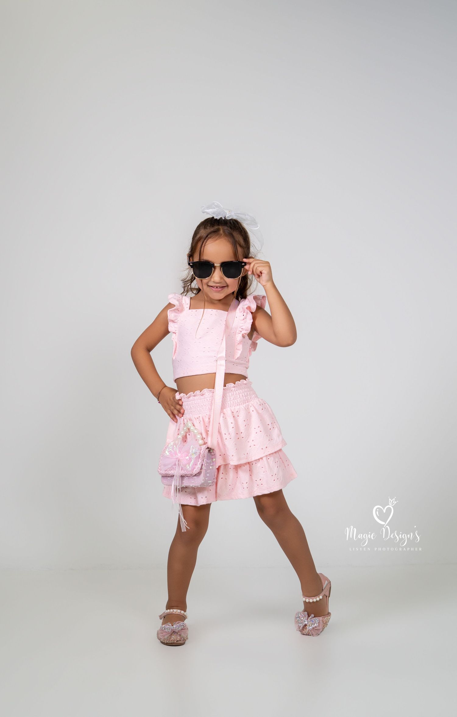 Child in pink ruffled outfit strikes playful poses with sunglasses against white background.
