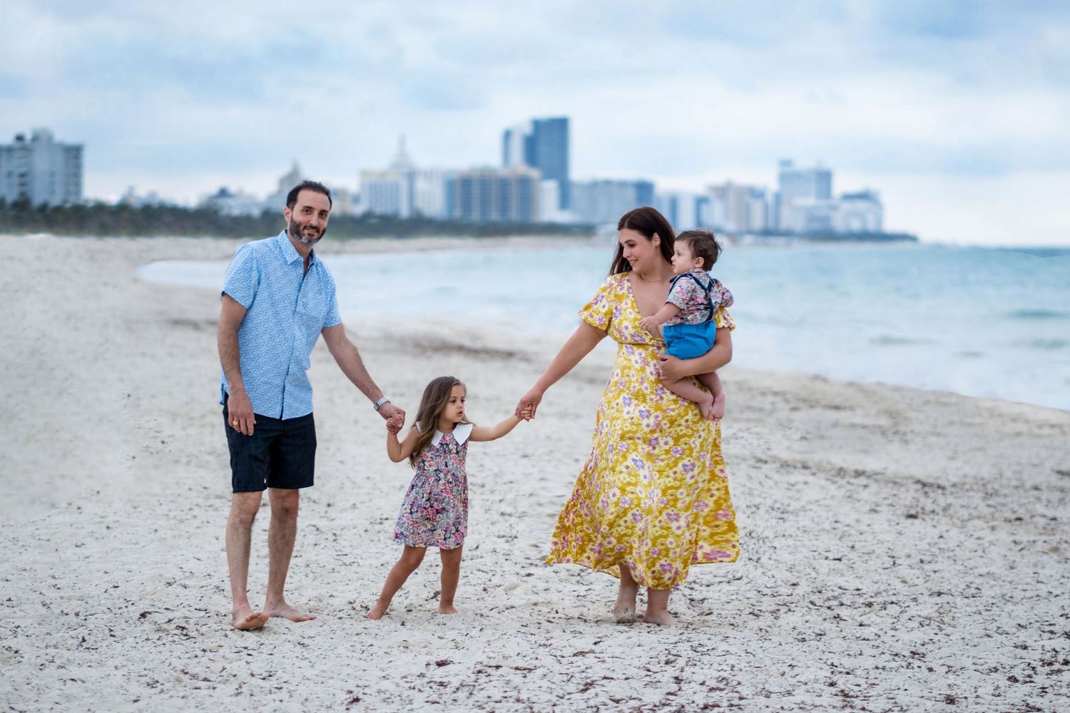 Family enjoying playful moments together on sandy beach with cityscape in background.