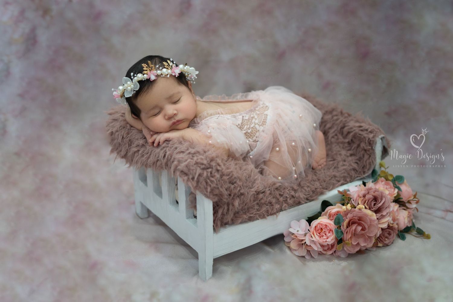Newborn photography setup with soft pink flowers and white miniature bed.
