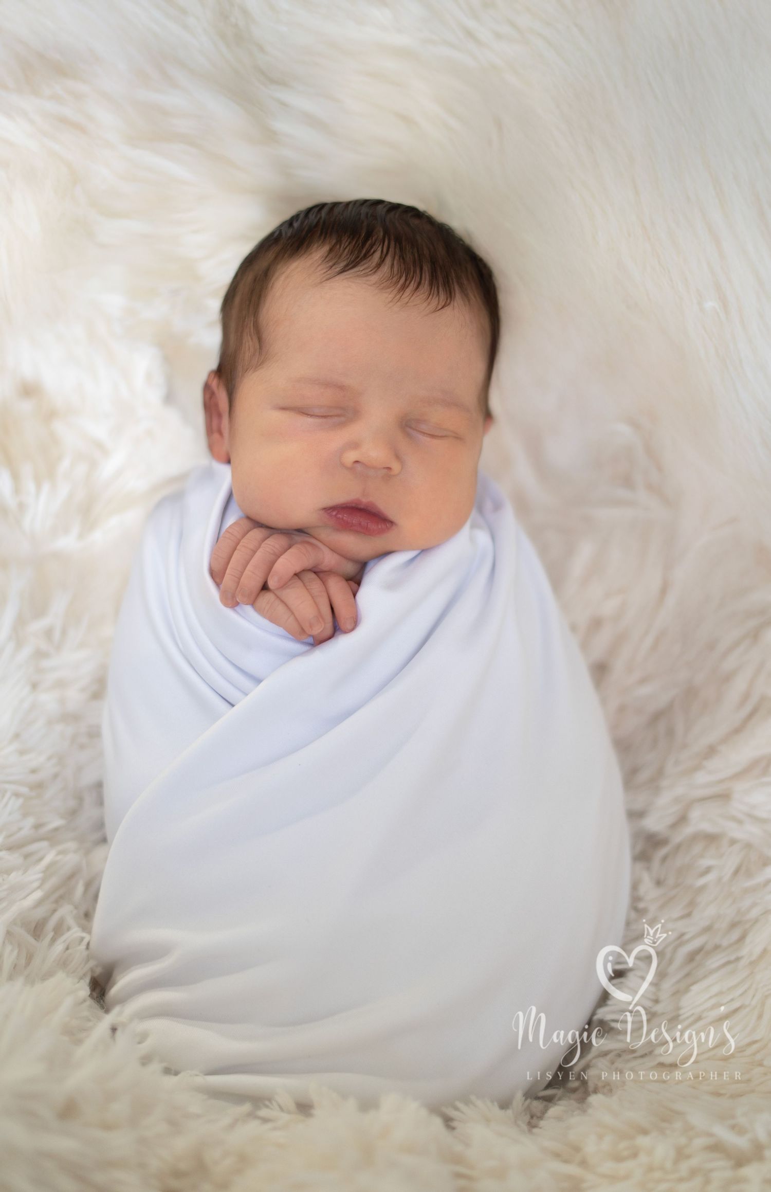 Newborn baby peacefully sleeping while wrapped in soft white blanket on furry white background.