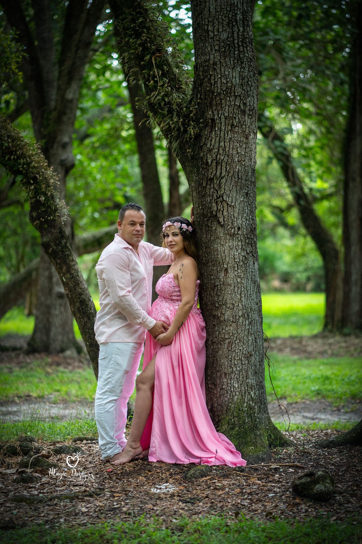 Romantic maternity photography session in a wooded area with pink dress and white shirt.