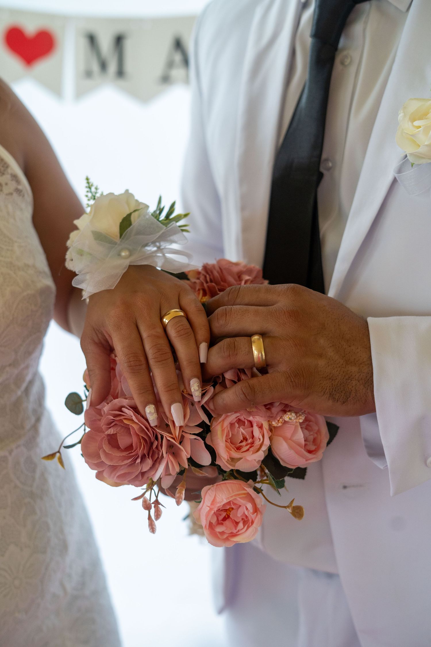 Close-up of newlyweds' hands with rings and pink rose corsage during wedding ceremony.