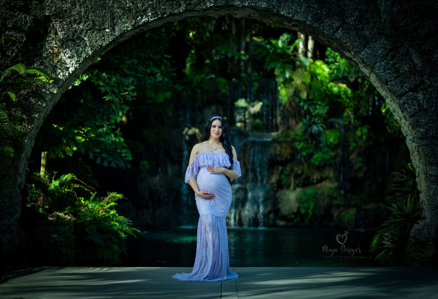 Elegant maternity portrait under stone archway wearing flowing blue gown.