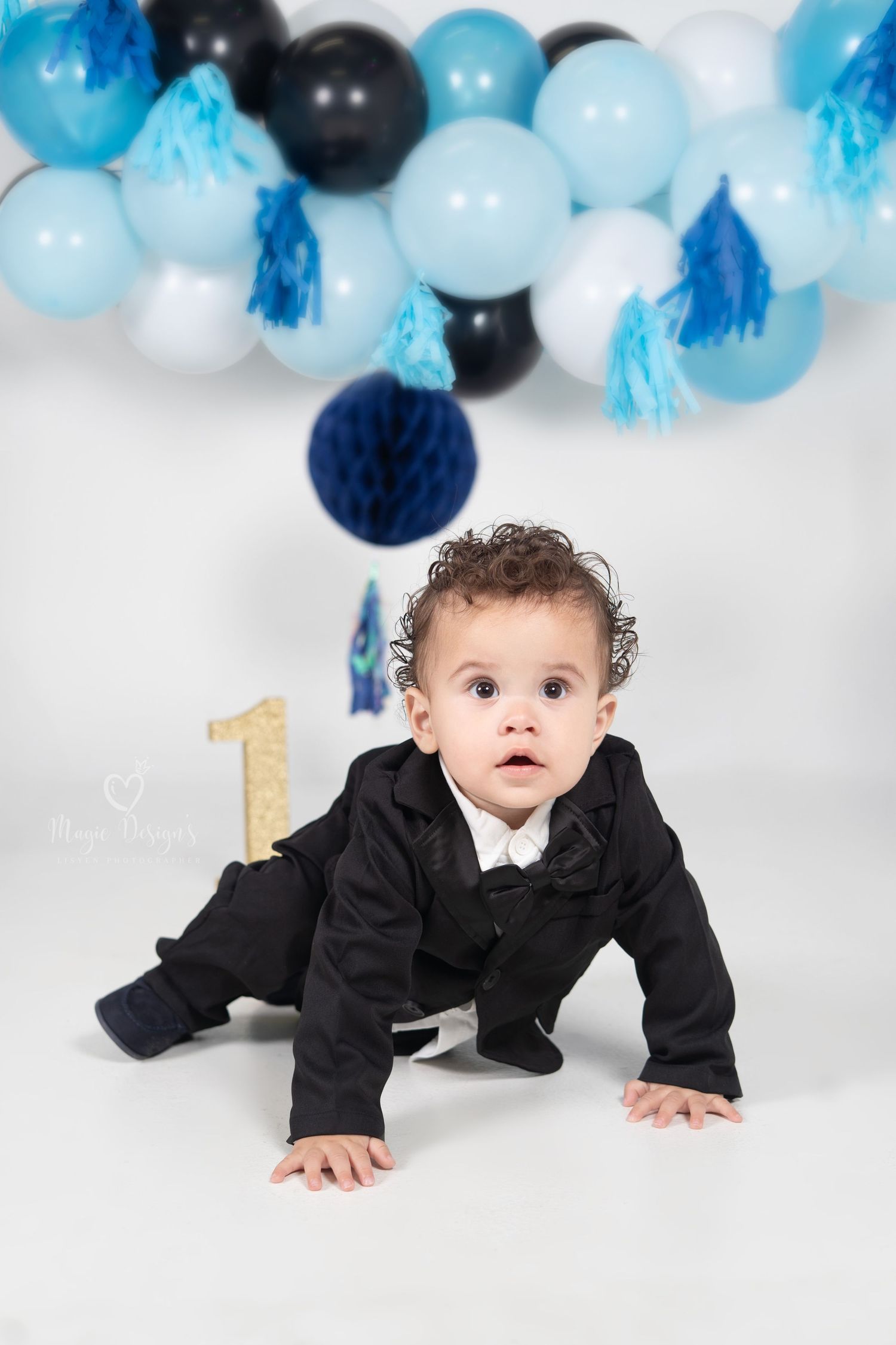 Baby in black suit crawls under blue and black balloon arch decorations for first birthday celebration.