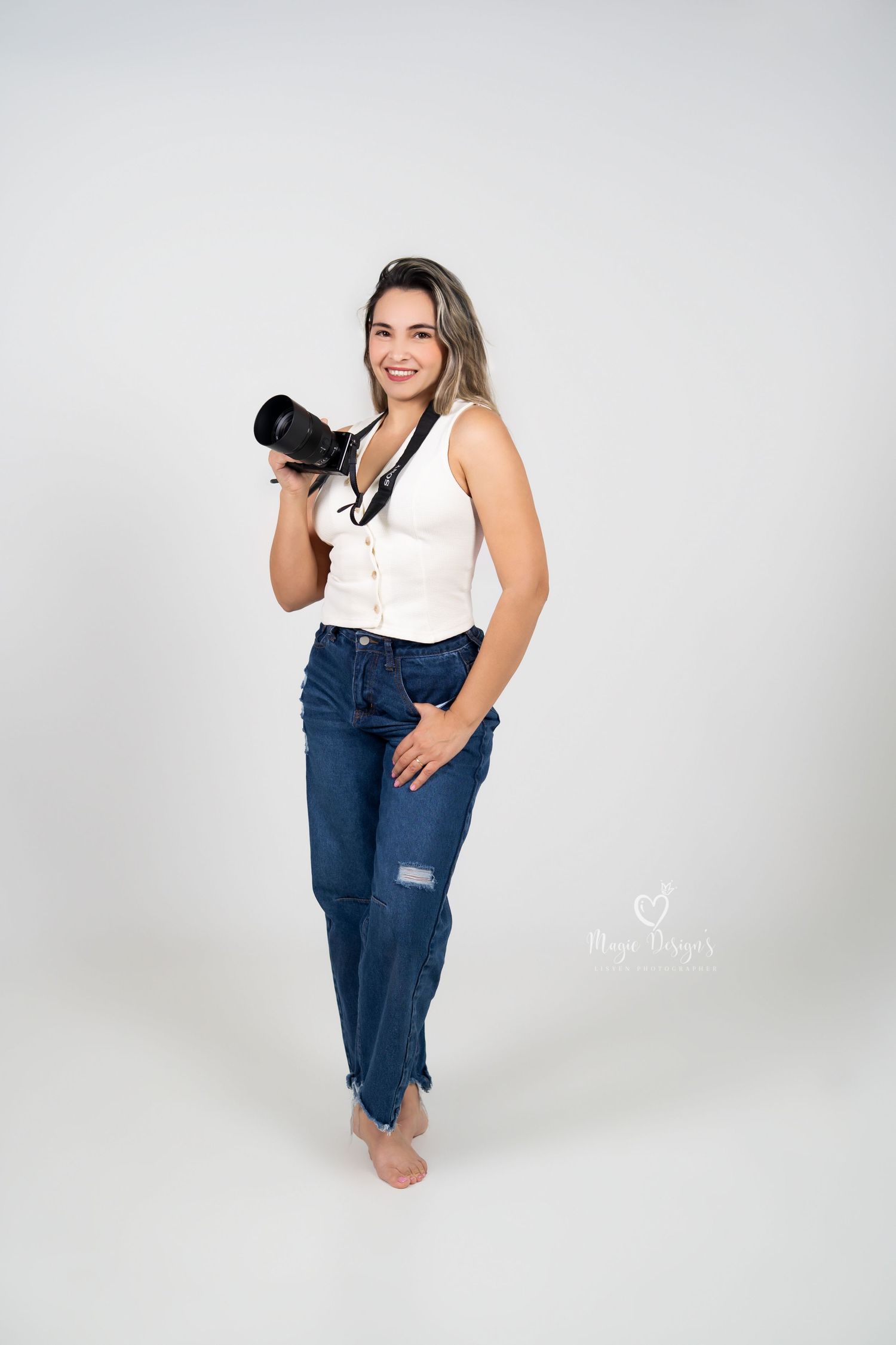 Professional photographer wearing white tank top and distressed jeans holds camera while posing barefoot in studio.
