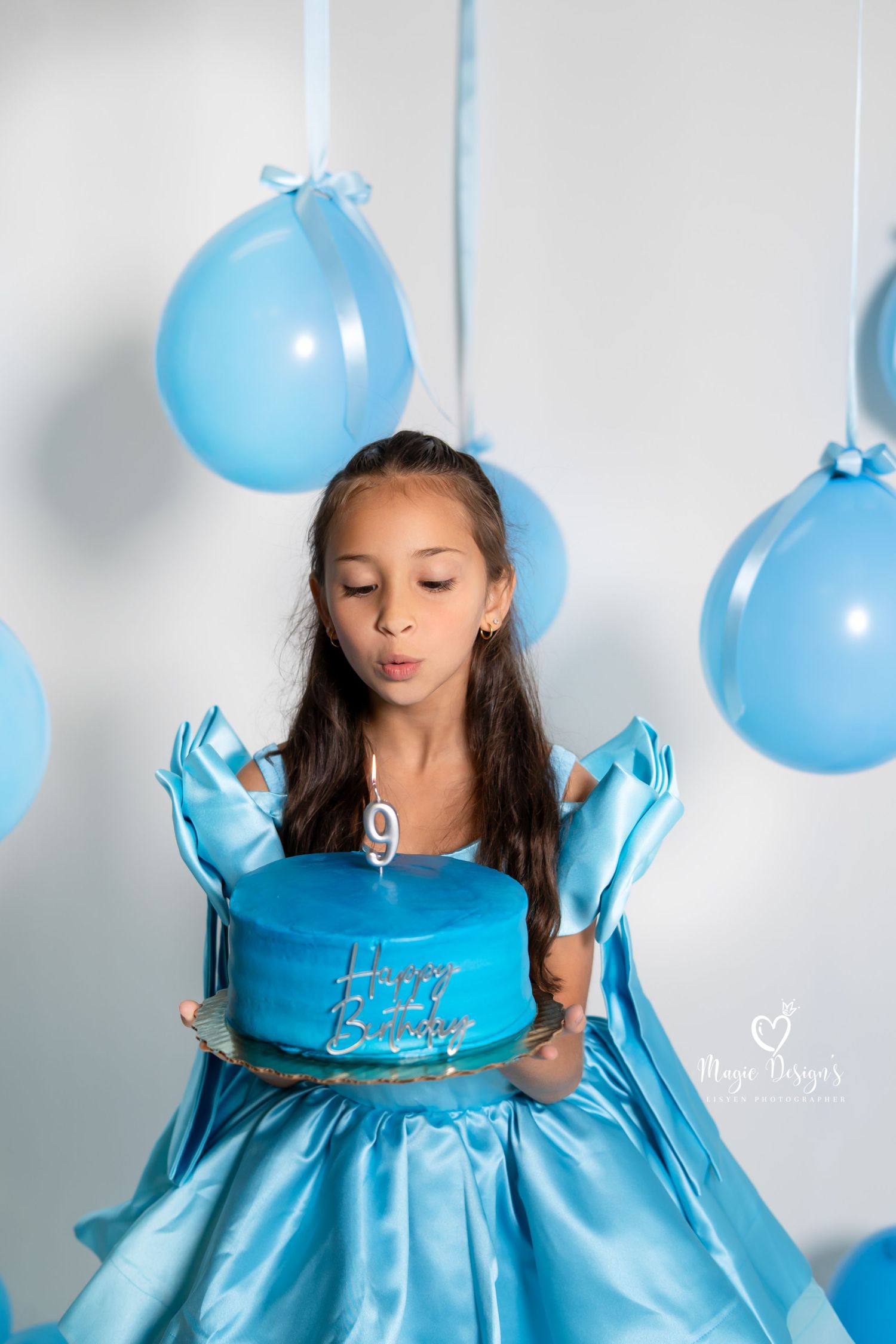 Young girl in blue dress poses with matching blue balloons against white wall in dreamy portrait series.