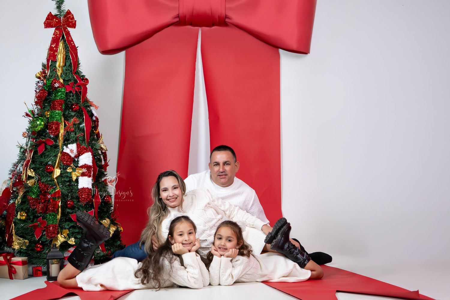 Family wearing white sweaters poses in front of Christmas tree with large red bow backdrop for holiday photos.