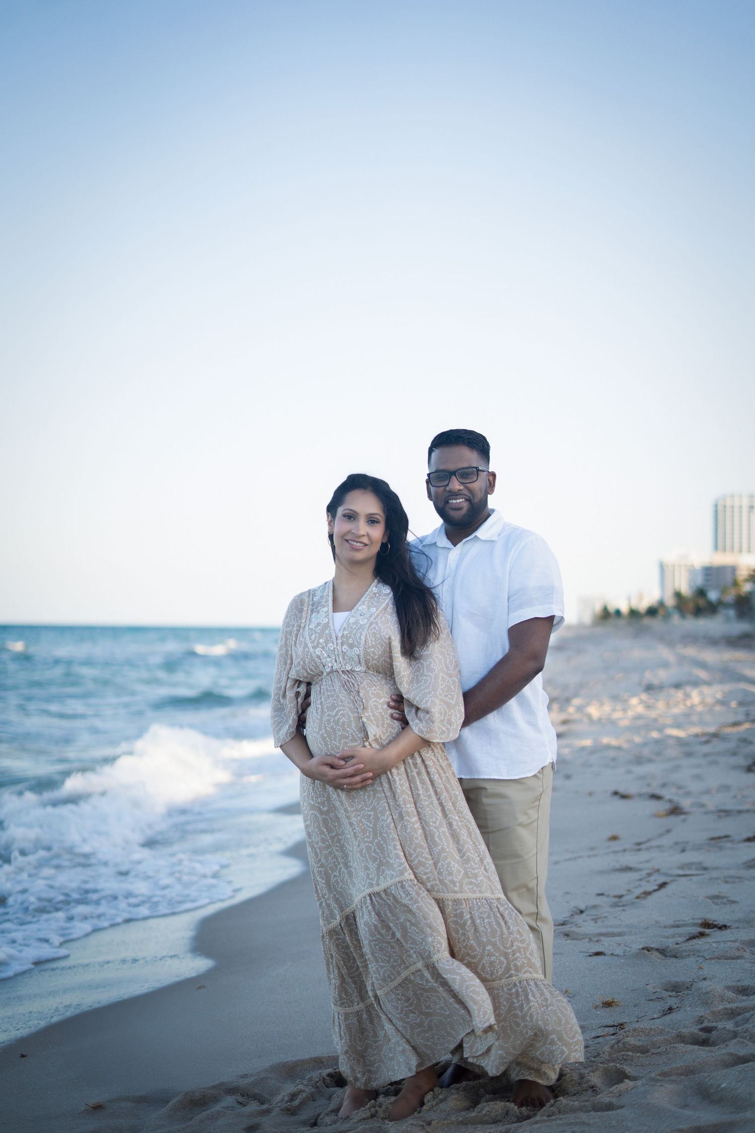 A couple shares a romantic moment on the beach at sunset wearing coordinated white and beige outfits.