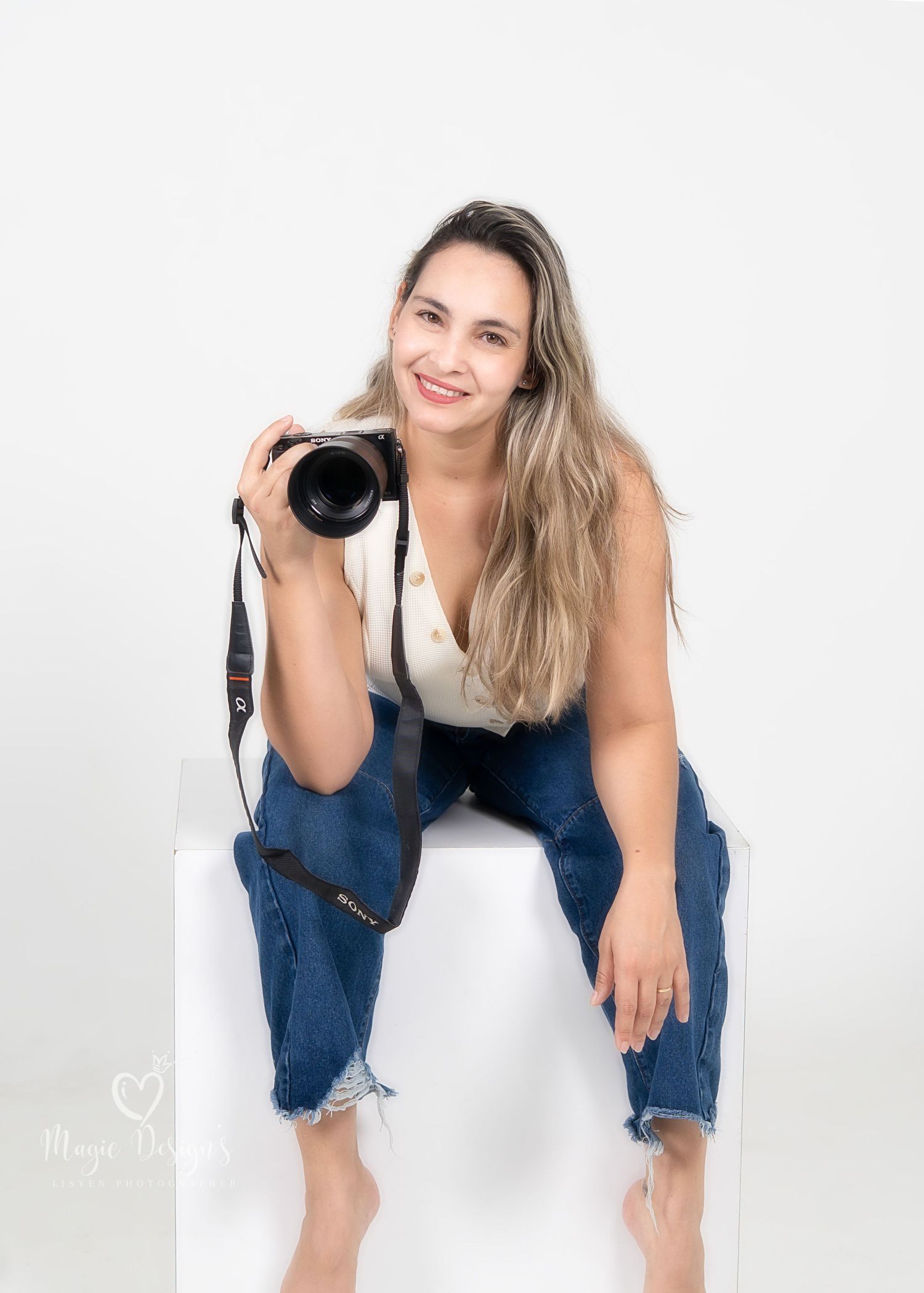 Professional photographer in distressed denim overalls poses with camera against white studio backdrop.