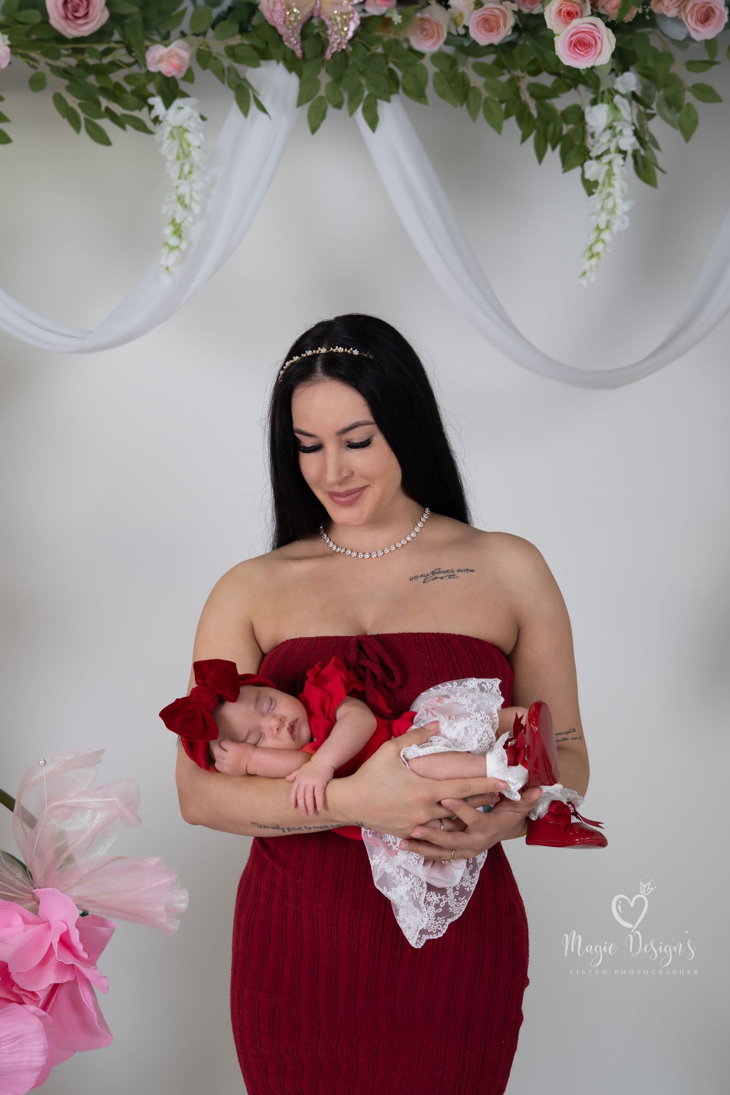 Mother and newborn share intimate moment under floral arch wearing red dress.