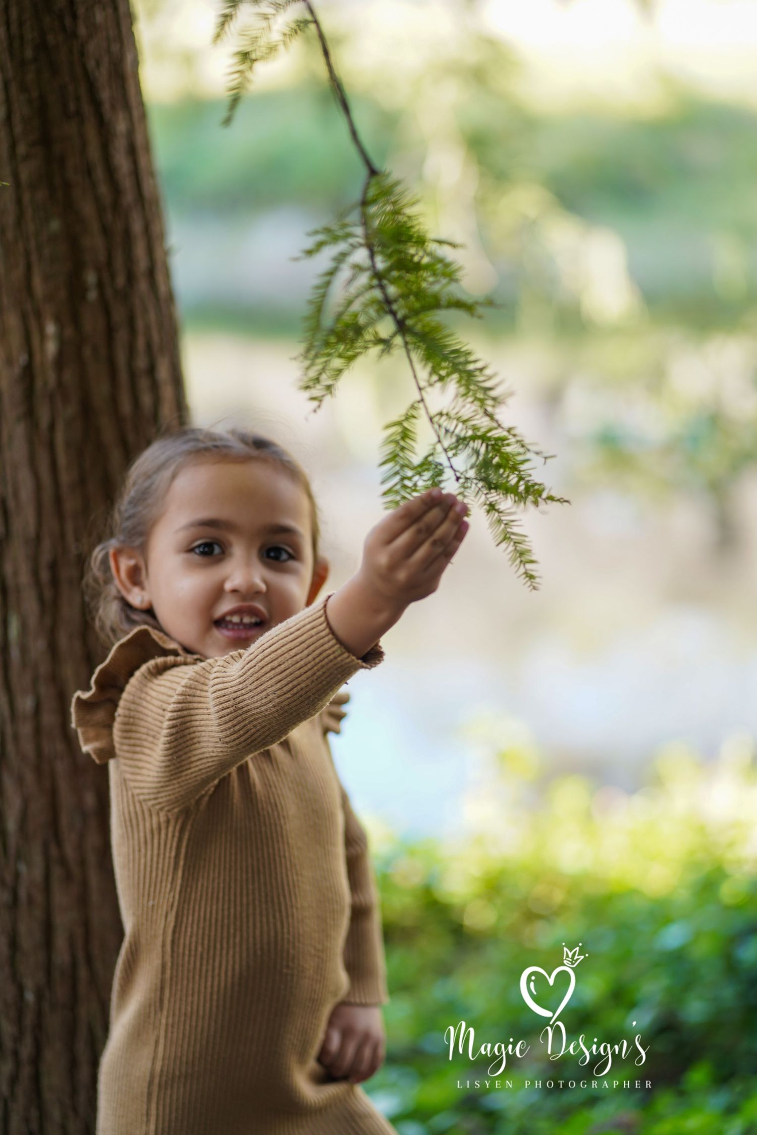 Child in brown sweater reaching up to touch tree branches in natural outdoor setting.