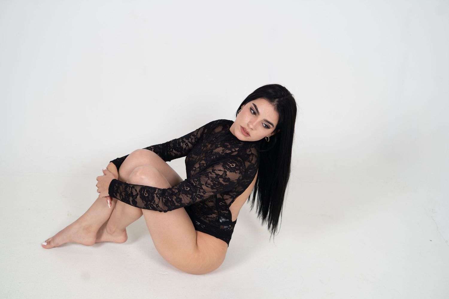 A figure in black lace bodysuit poses on white backdrop in studio setting.