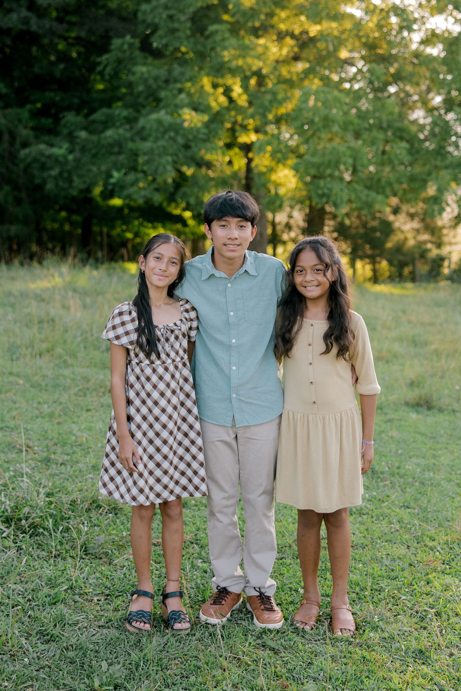 Three children pose together outdoors in a grassy field, wearing casual summer attire with trees in the background.