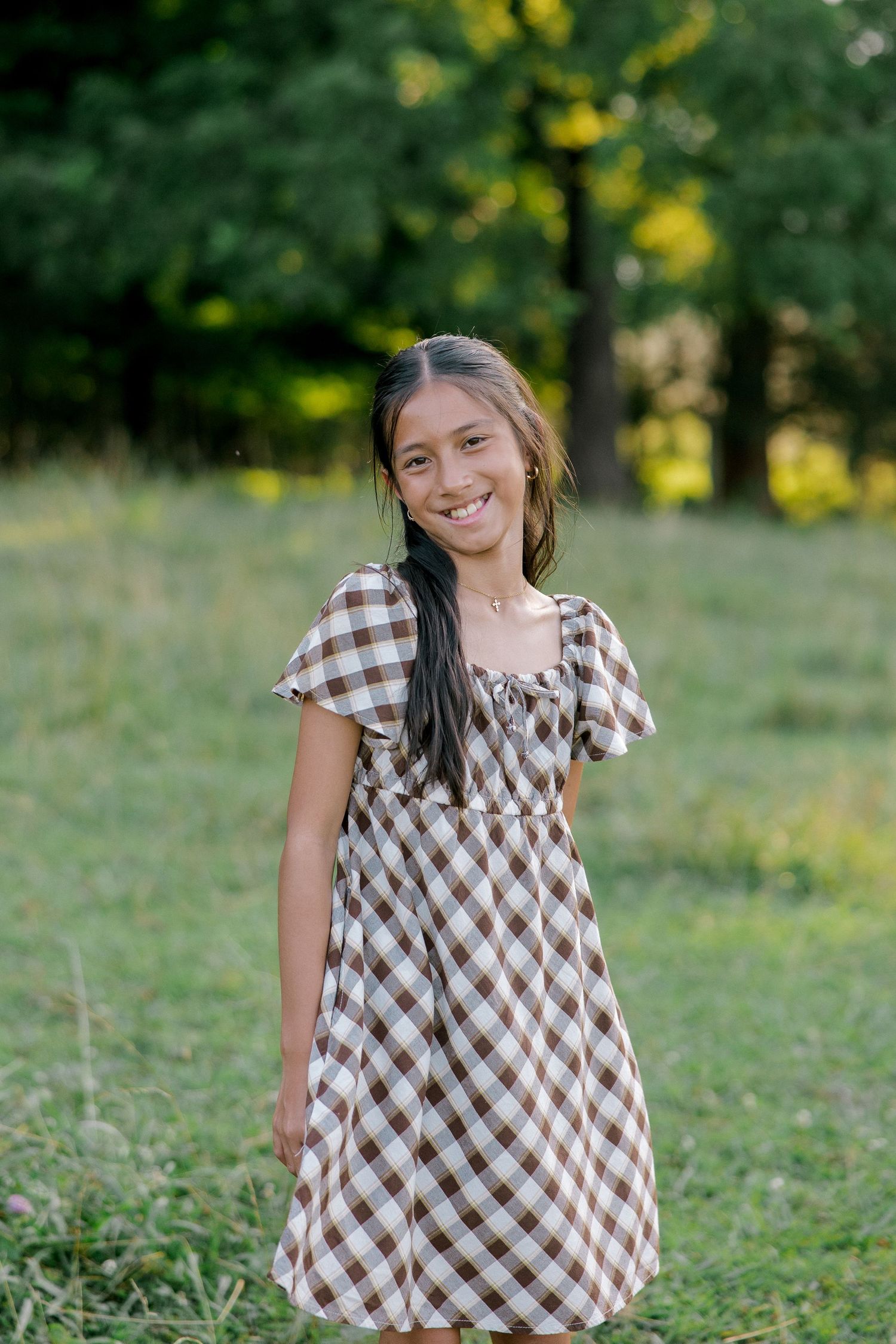 A smiling young girl in a brown and white gingham dress standing in a green park.