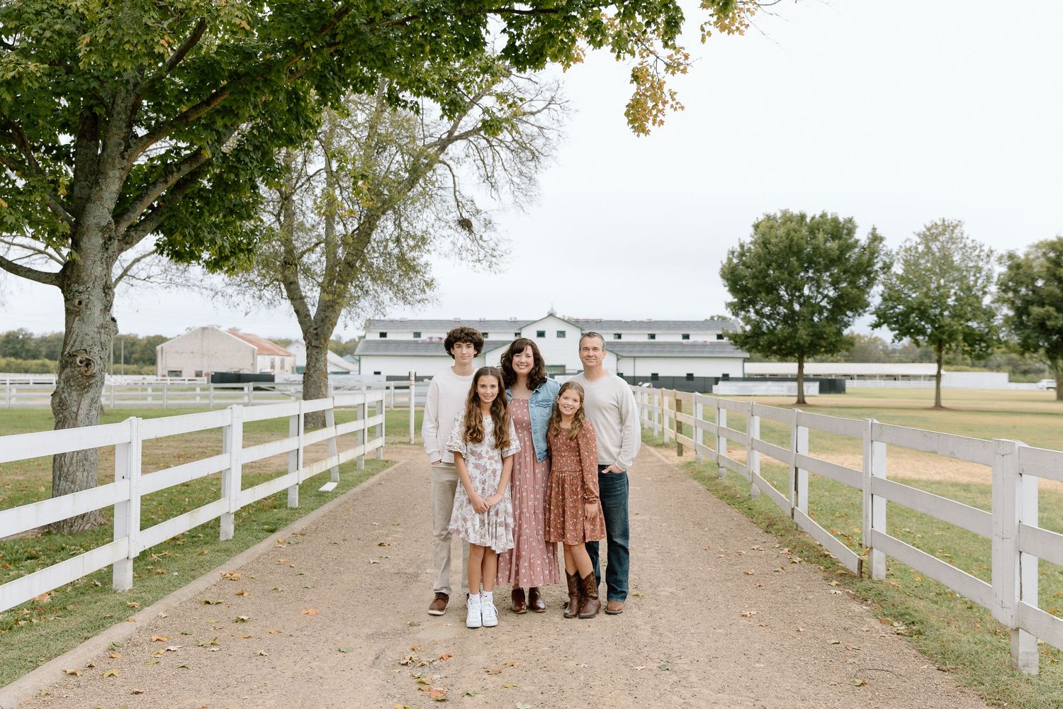 Family of five posing on a tree-lined gravel path by white fence at a farm.