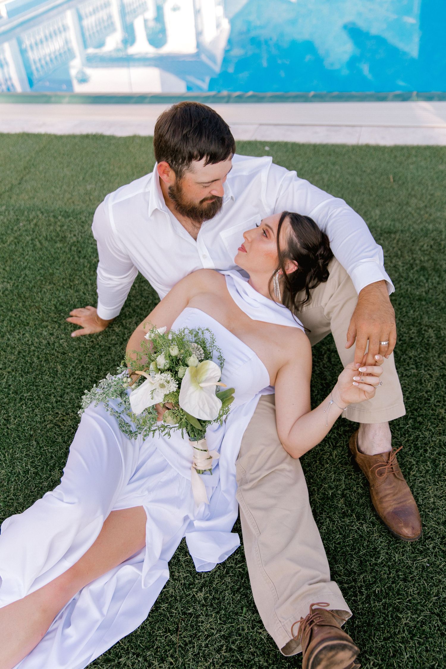 Bride and groom sitting on green grass near pool, bride holding white bouquet in wedding dress, groom in white shirt.