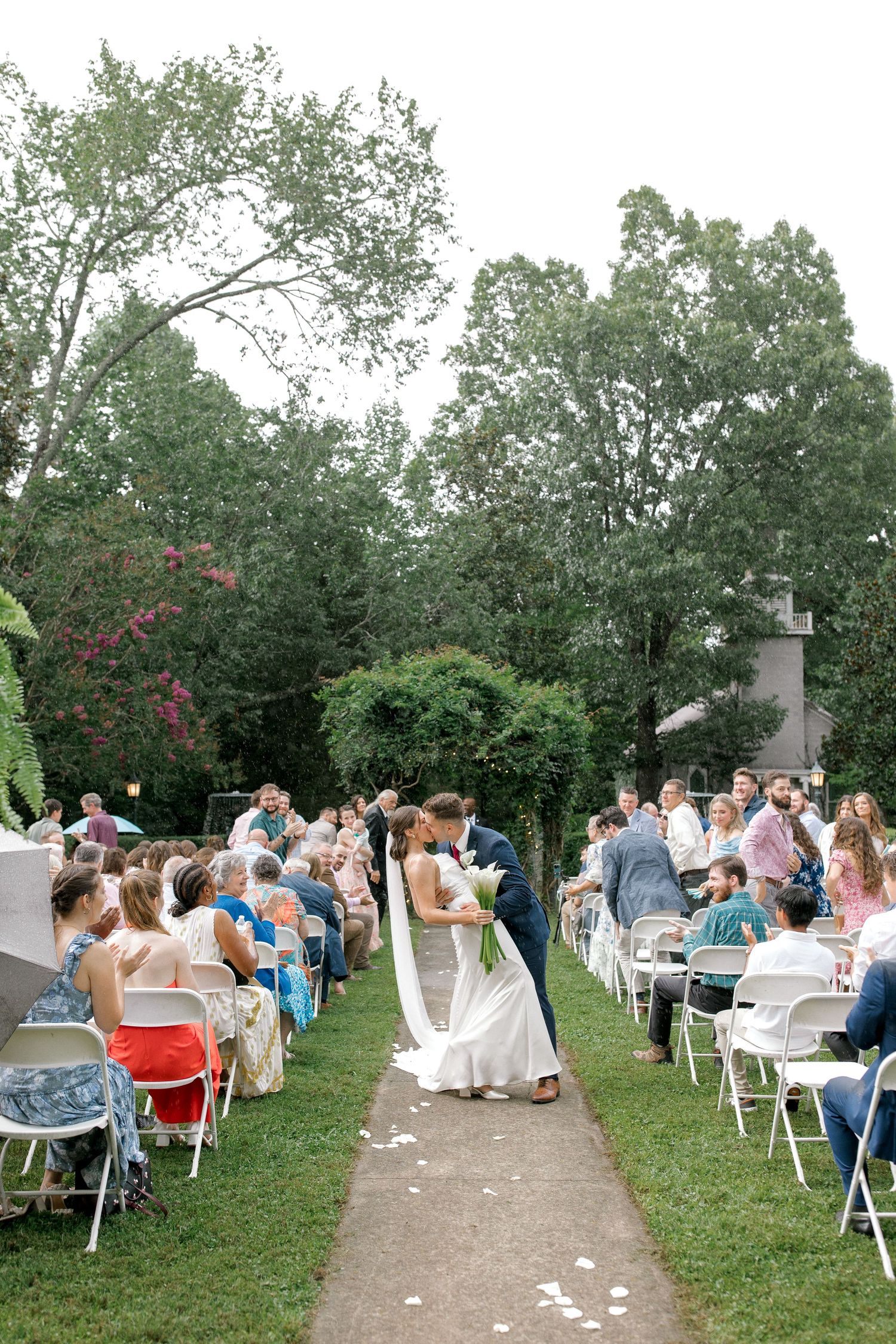 Bride and groom share first kiss at outdoor wedding ceremony aisle surrounded by seated guests.