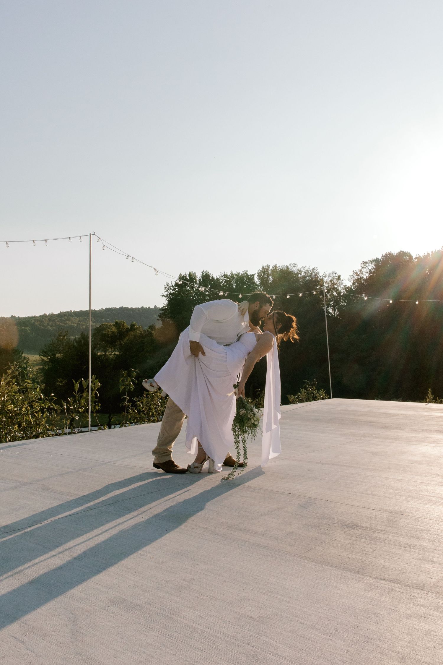 A couple shares a romantic dip kiss on an outdoor dance floor at sunset during their wedding celebration.