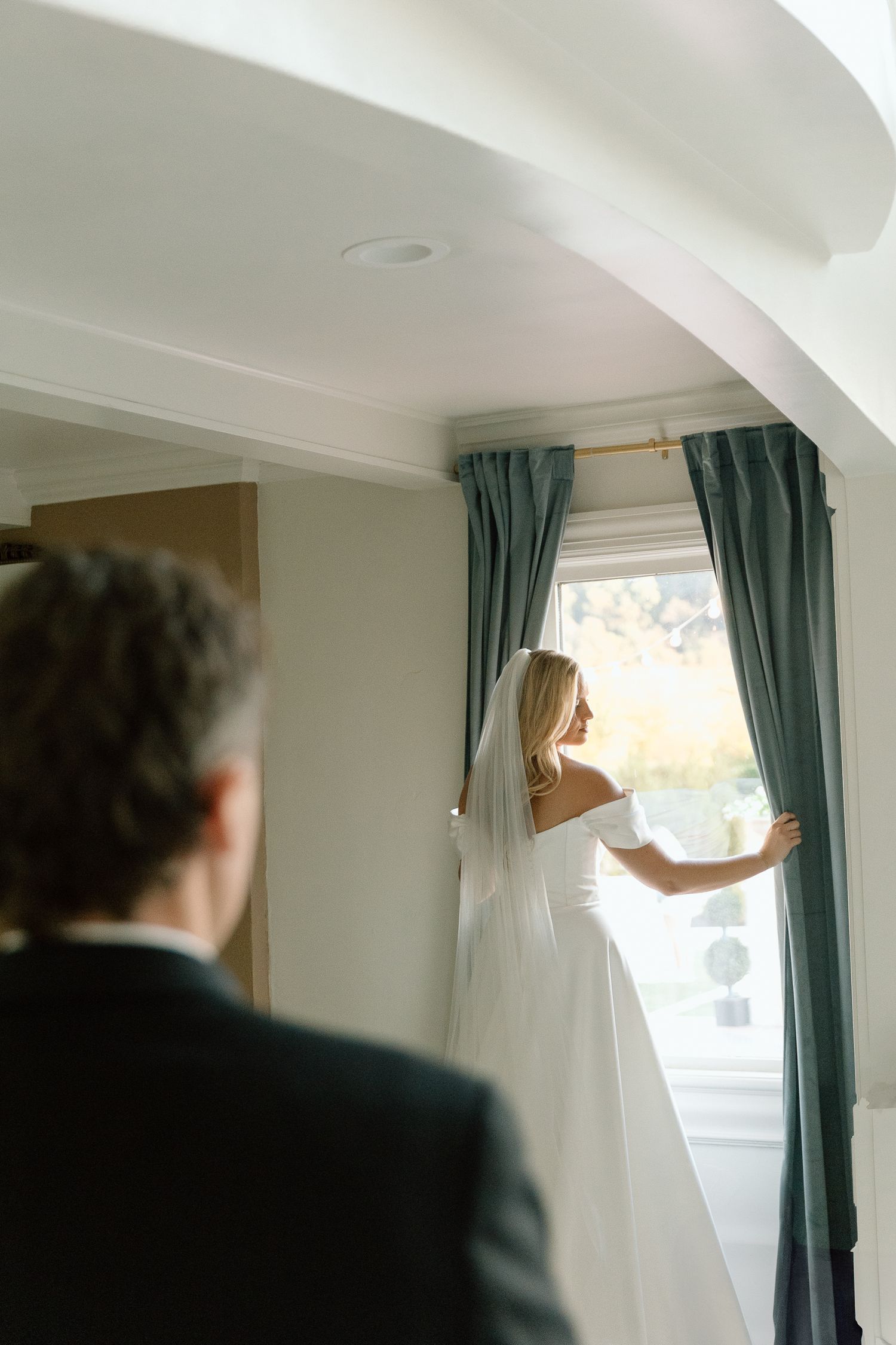 Bride in white gown and veil stands at window with blue curtains as groom looks on in elegant wedding venue.