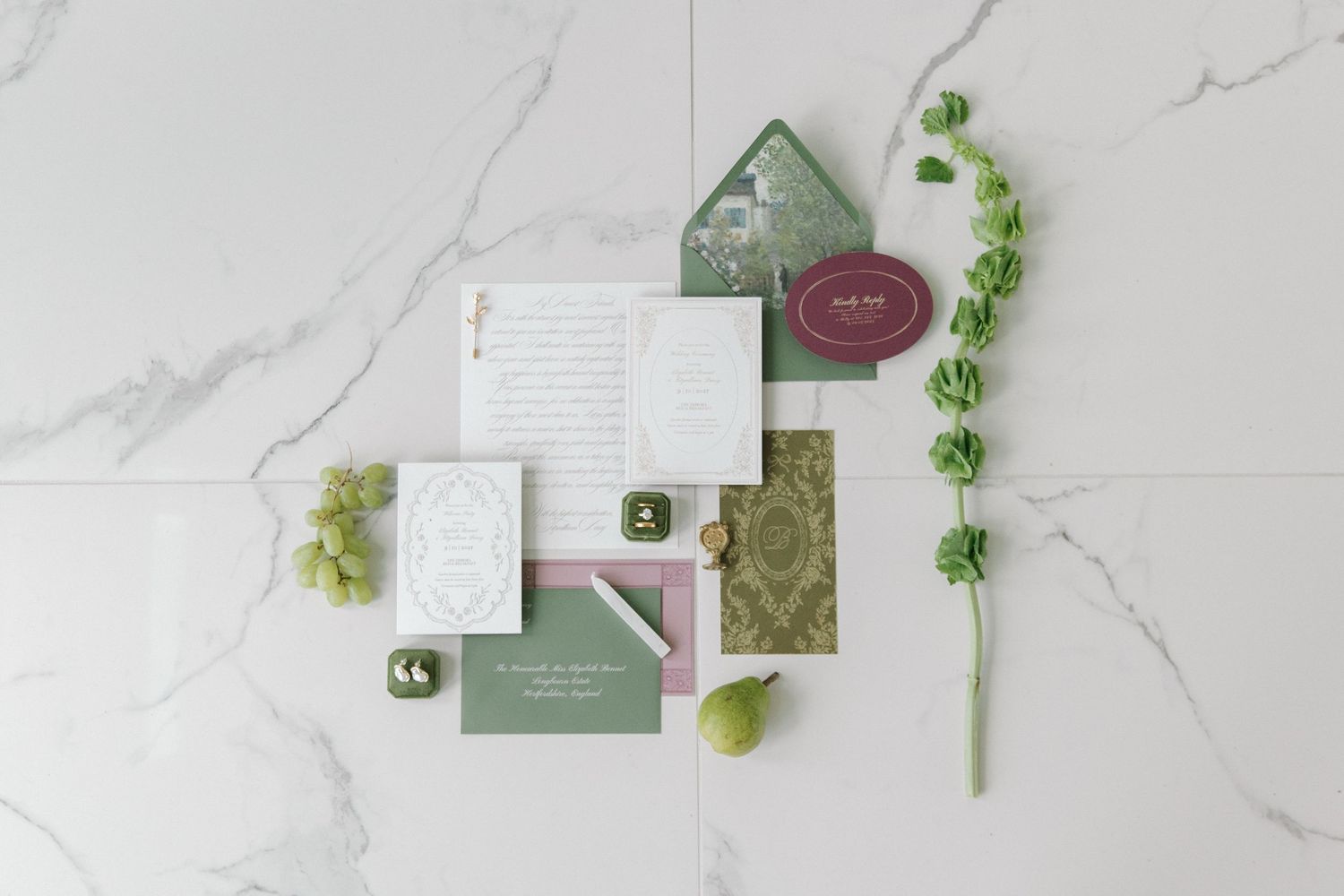 Elegant wedding stationery flatlay with green and burgundy invitations, wax seal, and floral accents on marble.