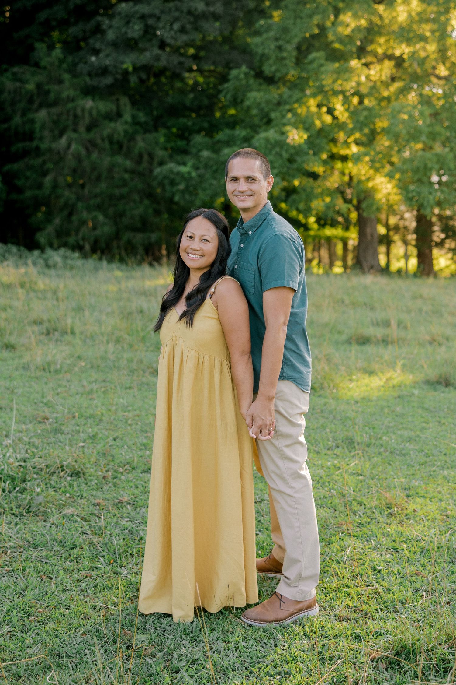 A smiling couple holds hands back-to-back in a sunny green meadow, woman in yellow maxi dress, man in teal polo shirt.