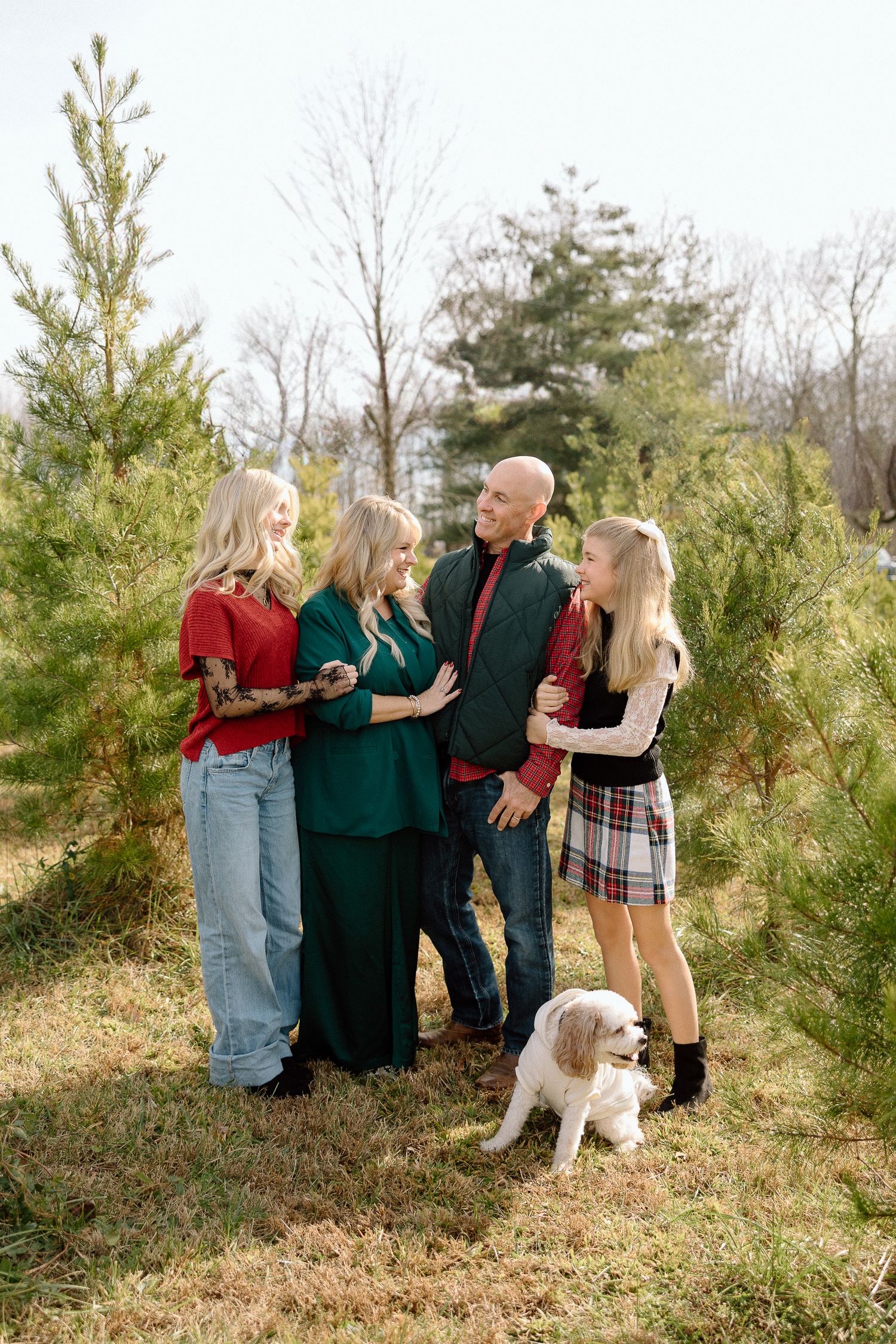 Family of four with a white dog posing among pine trees outdoors, wearing festive red and green holiday outfits.